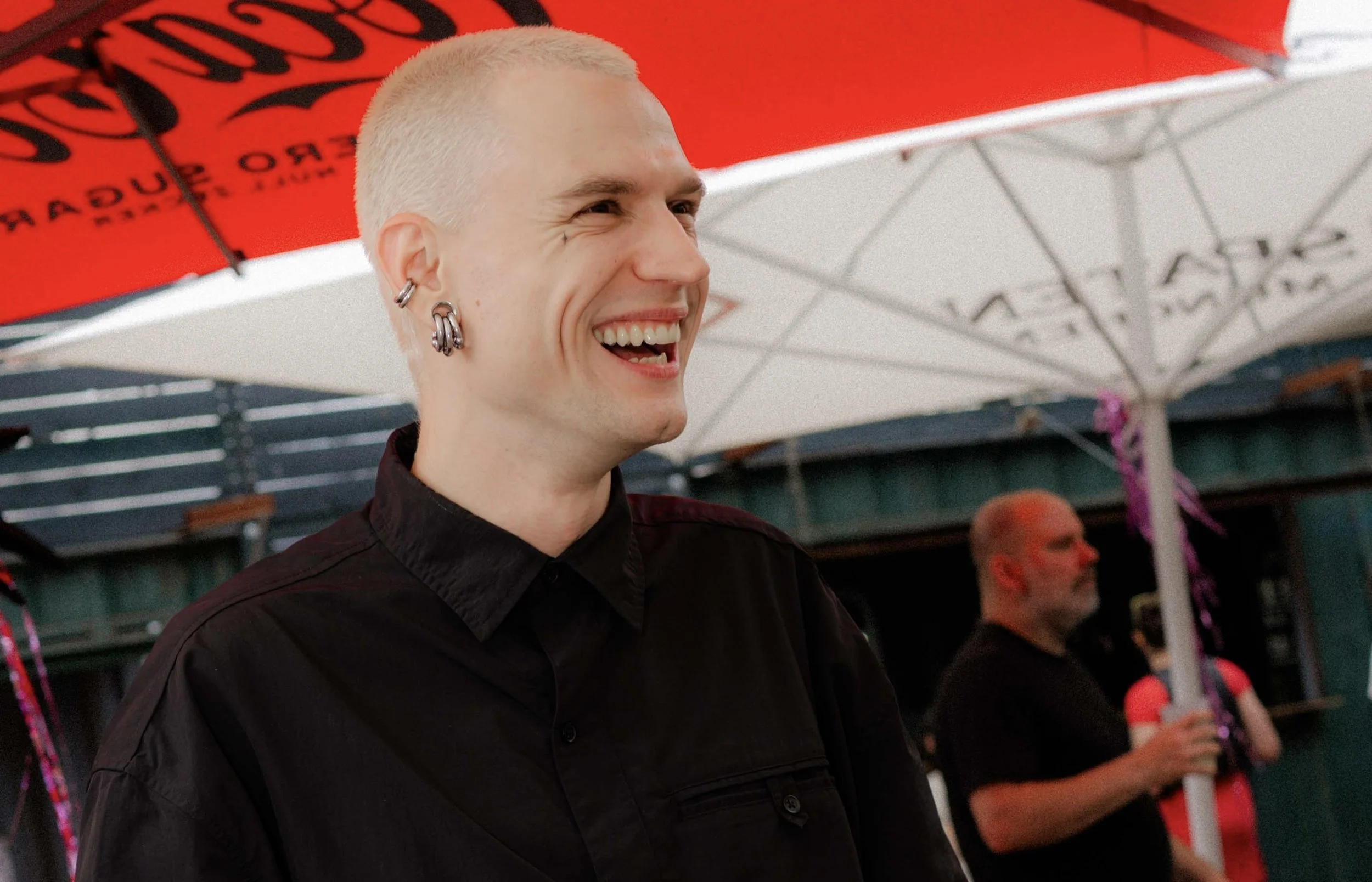 A person with short bleached hair and multiple earrings smiling under a red umbrella.