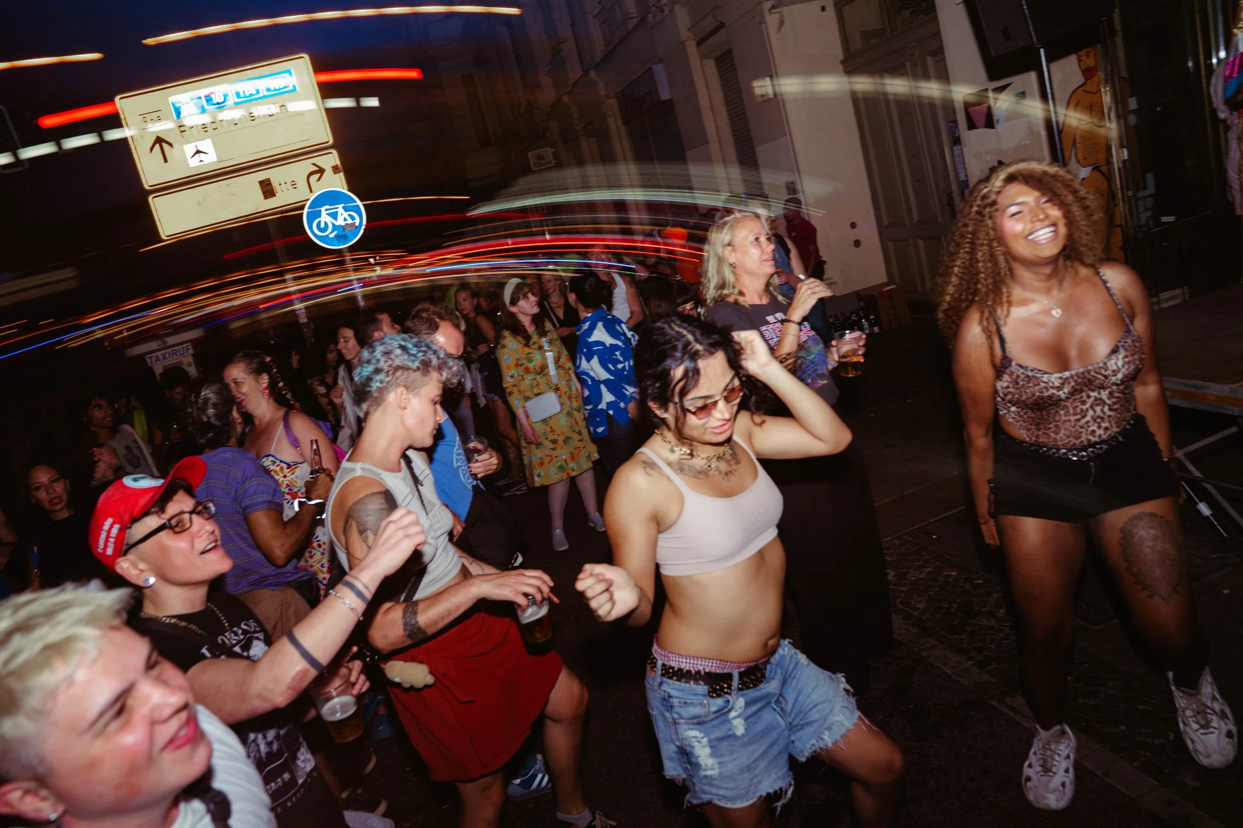 People dancing and having fun at a lively night event on a city street, with some smiling and enjoying the music, illuminated by colorful lights.