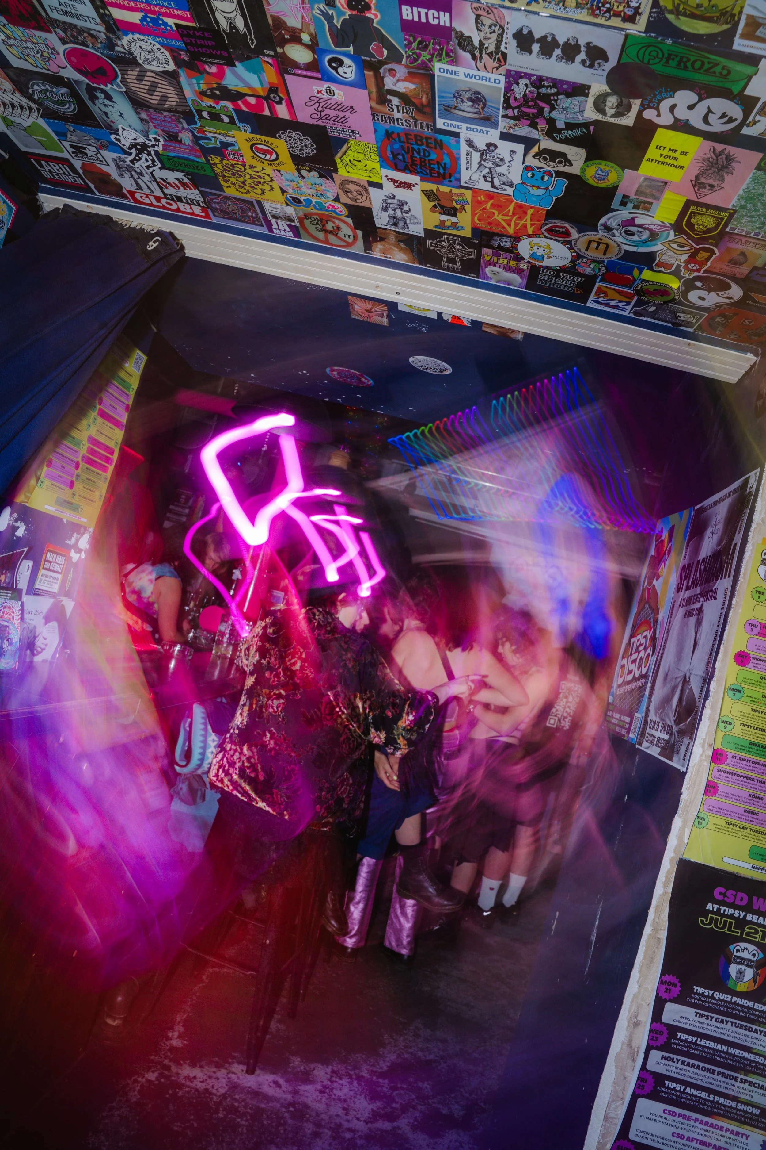 A wall covered in numerous colorful stickers and posters, with some neon pink and purple lighting reflections and a reflection of people standing in front of it.