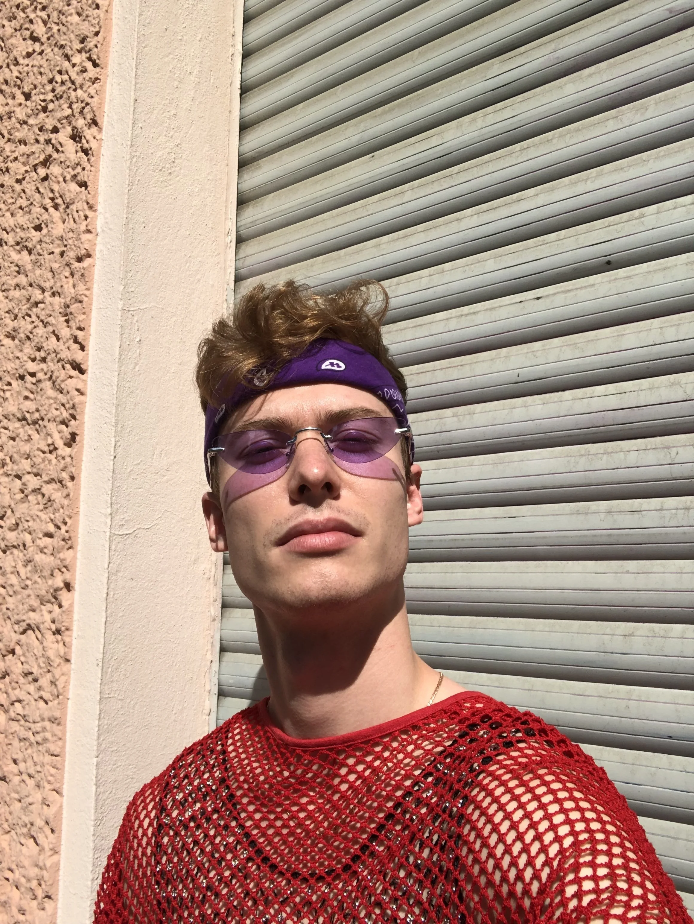 Close-up of a young person wearing purple-tinted sunglasses, a purple bandana, and a red netted top, standing outside against a background of a textured beige wall and a gray metal shutter.