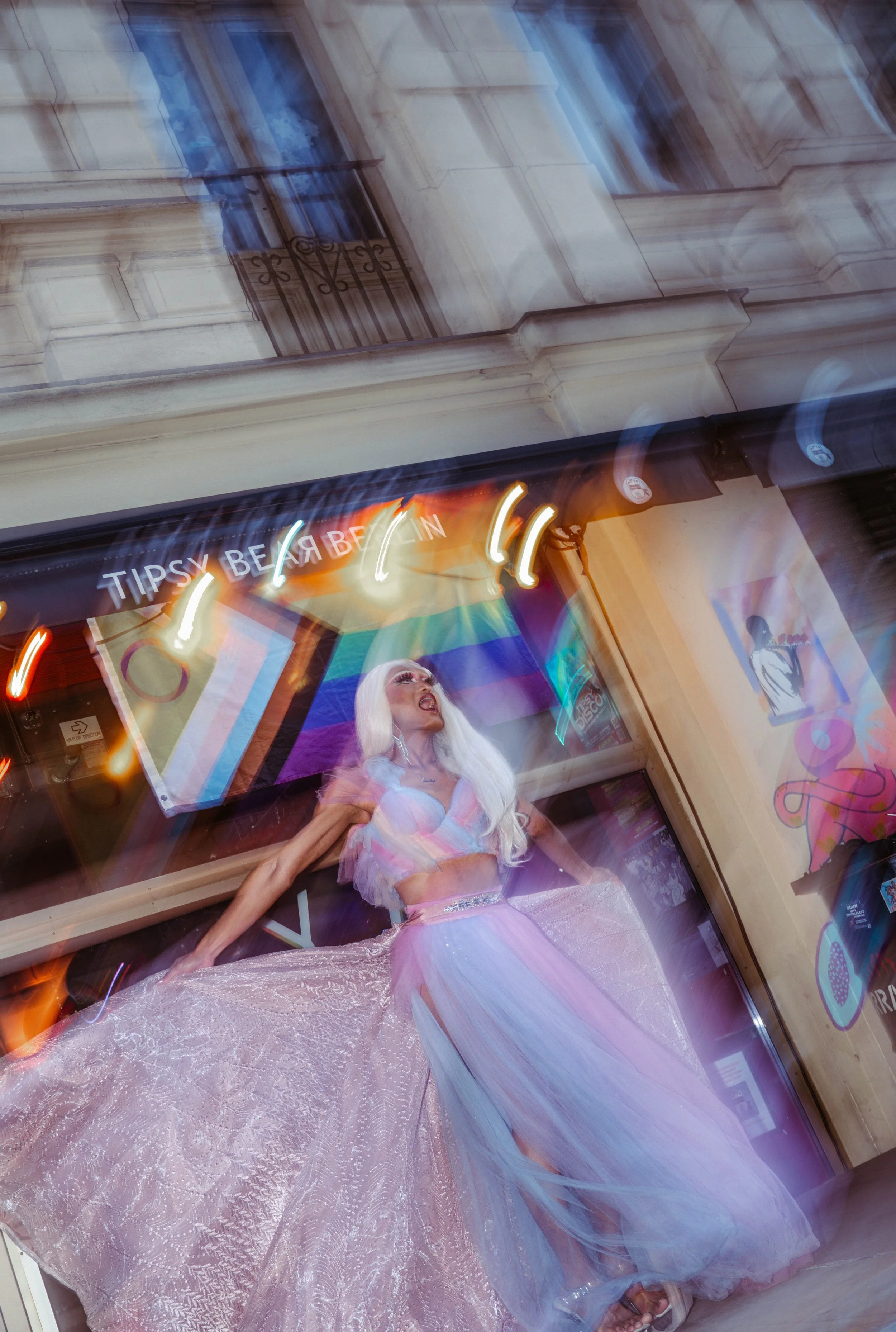 A woman with long blonde hair, wearing a pastel pink ball gown with shiny fabric and a sheer tulle overlay, posing in a store window display. The display has colorful neon signs and a sign that says "Tipsy Beverly". The background reflects a building