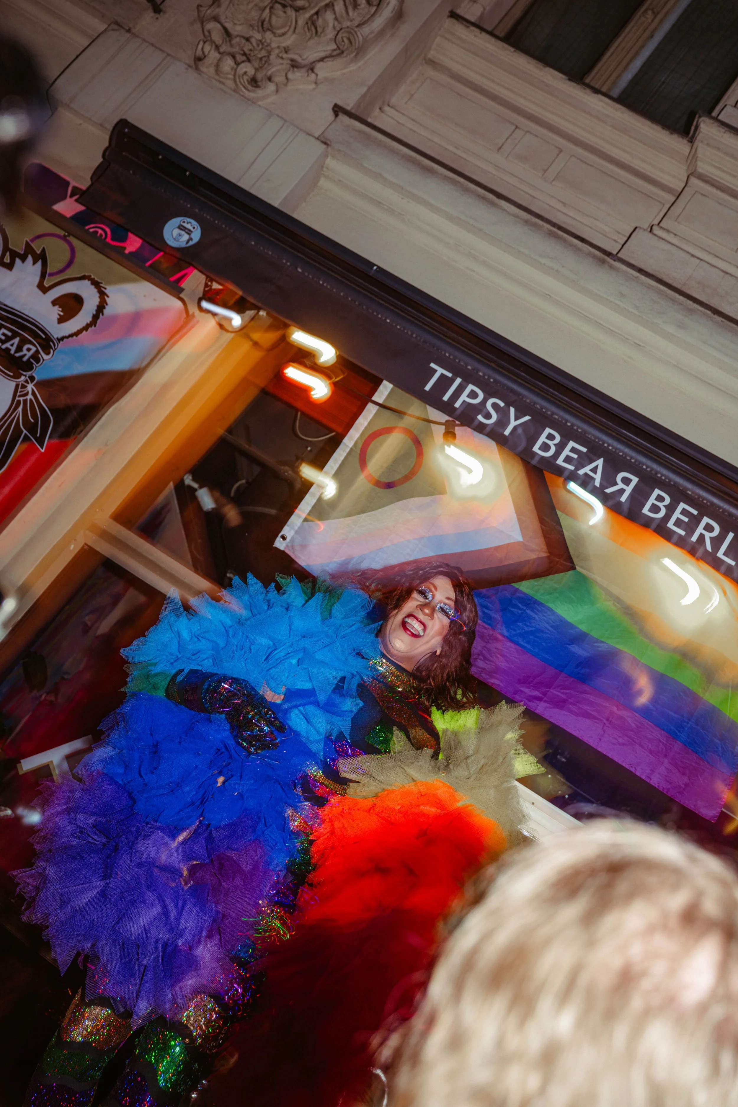 A person dressed in a multicolored, feathered costume, smiling, standing in front of a rainbow flag, with a sign overhead reading 'Tipsy Bear.'