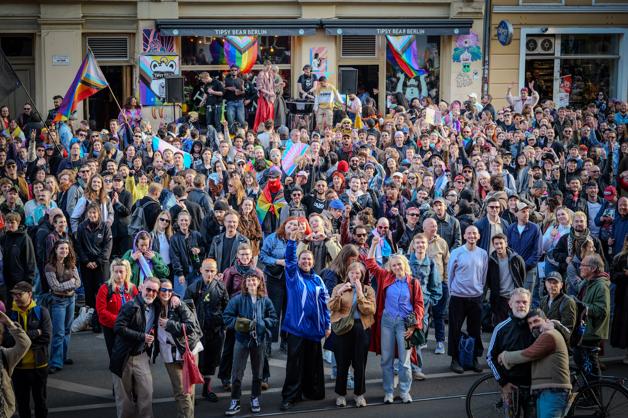 A large crowd of people gathered outdoors in front of a building, celebrating at a pride event. Some are holding rainbow flags and wearing colorful clothing. There is a raised stage with DJs and performers behind the crowd, and colorful artwork and flags decorate the scene.