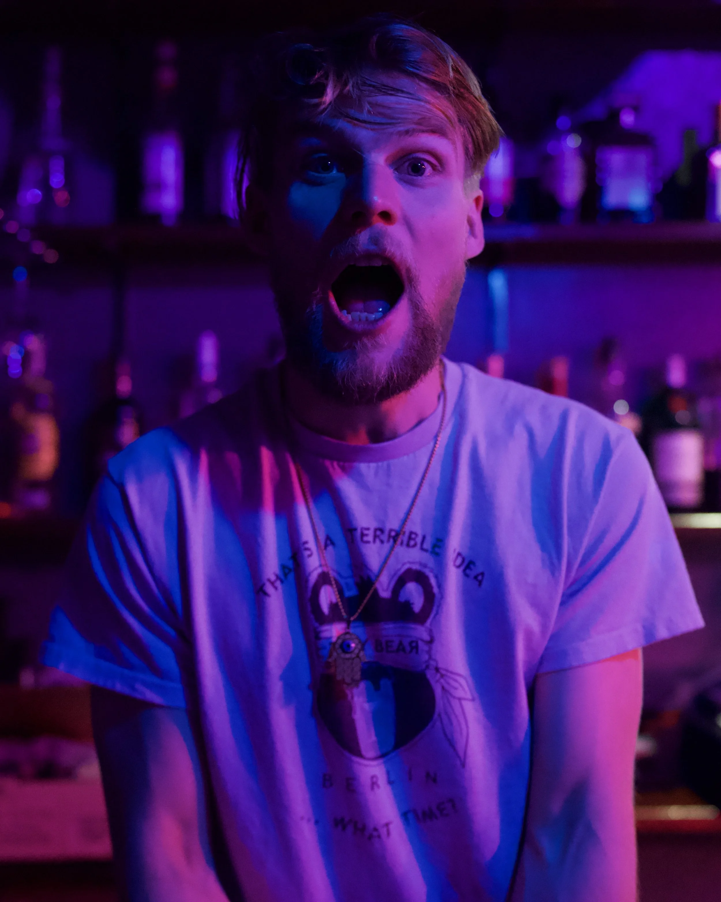 A man with a surprised or shocked facial expression, mouth open, in a dimly lit setting with purple and pink lighting, shelves of bottles in the background.