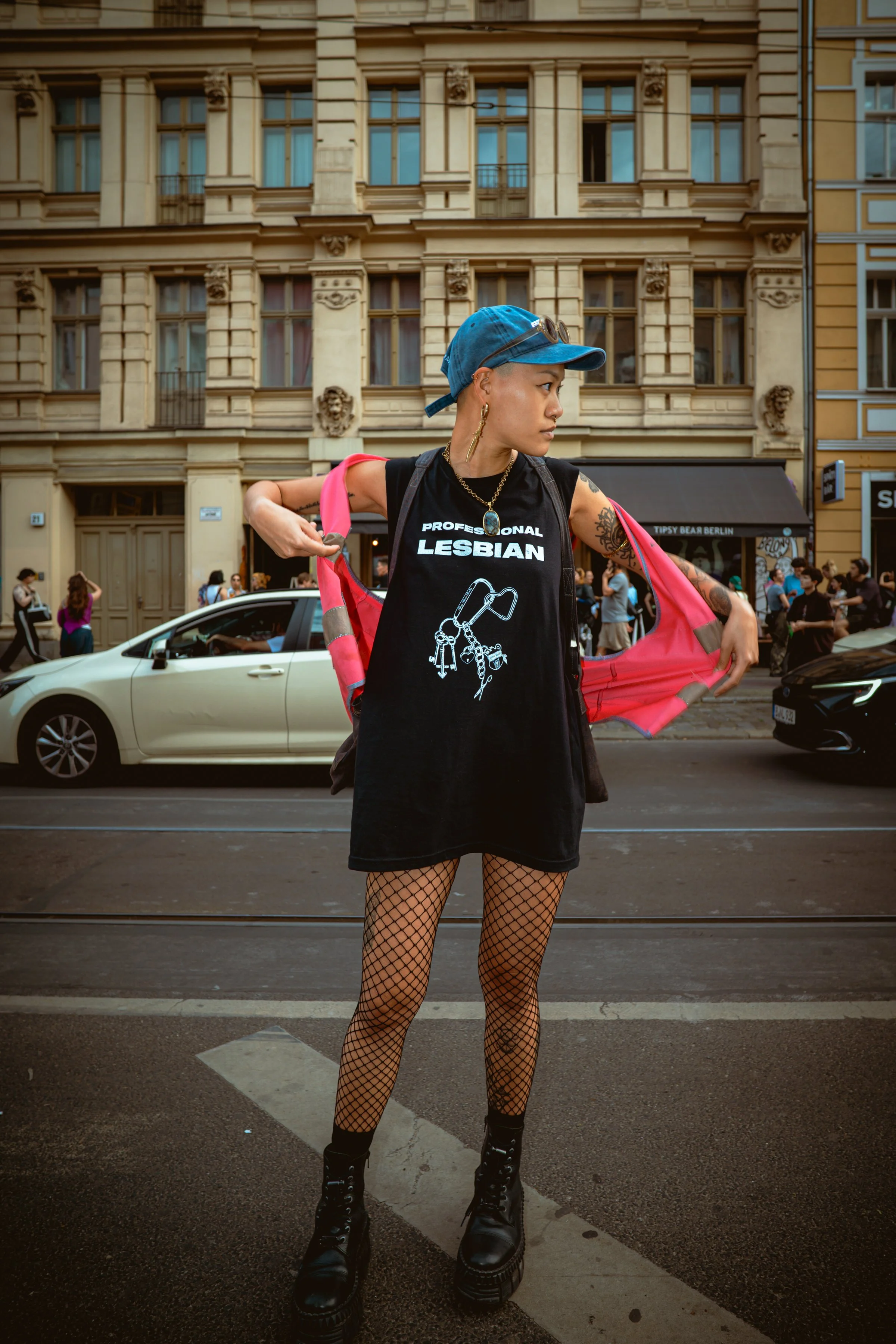 A person with short hair wearing a blue baseball cap, black sleeveless shirt with the words 'Professional Lesbian' and a graphic of handcuffs, fishnet stockings, black combat boots, and gold jewelry, standing on a city street with motorcycles, cars, pedestrians, and historic buildings in the background.