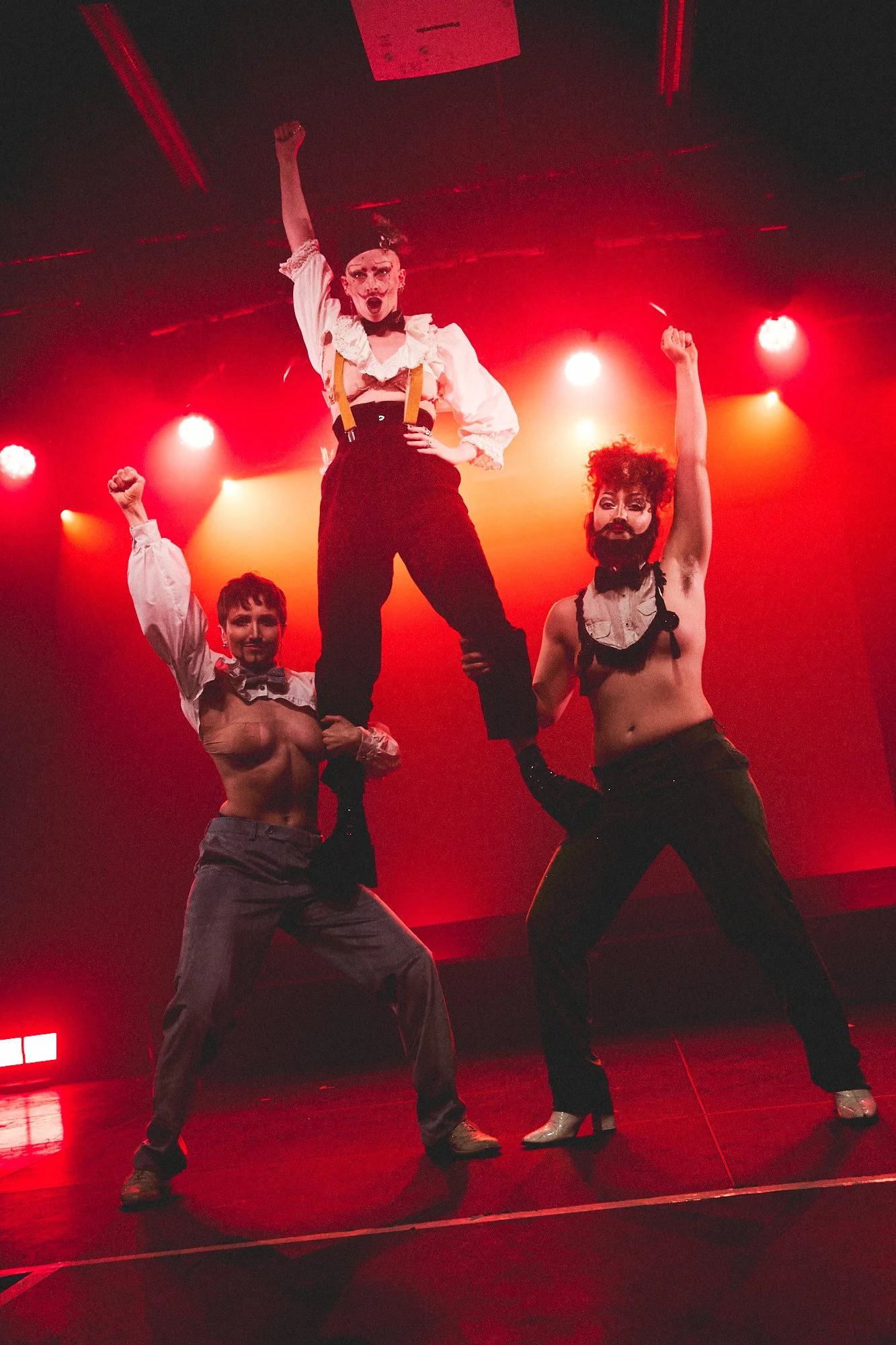 Three performers on stage under red lighting, with one person standing on the shoulders of another, both raising their fists, and the third person standing beside them with a raised fist, dressed in a theatrical and expressive style.