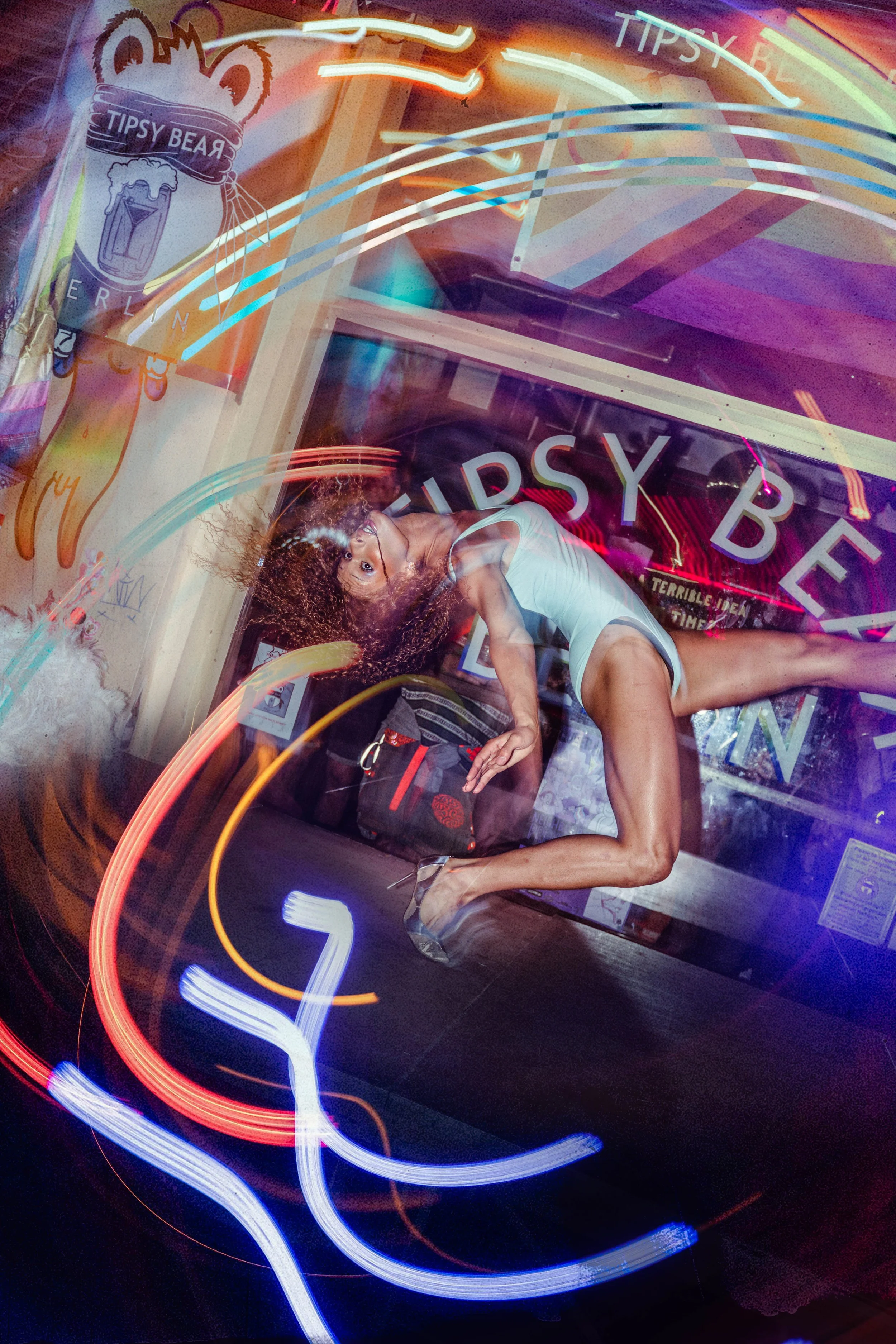 A woman with curly hair wearing a white tank top and shorts in a dance pose inside a room illuminated by colorful neon lights and signs, including one that says 'Tipsy Bear.' The image is overlaid with abstract light trails.