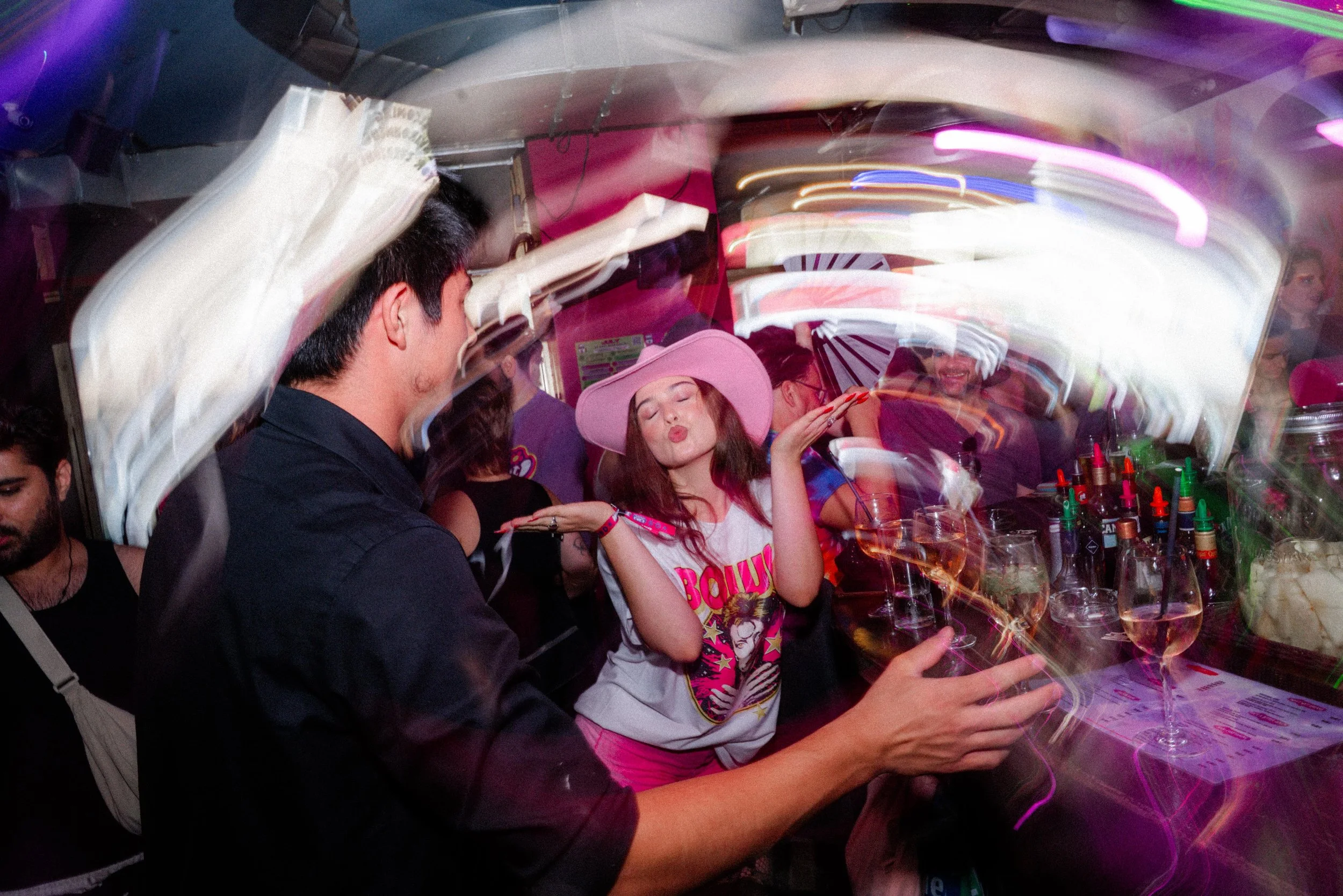 People at a party or bar, with a girl wearing a large pink hat, making a kissing face, surrounded by drinks and colorful lights.