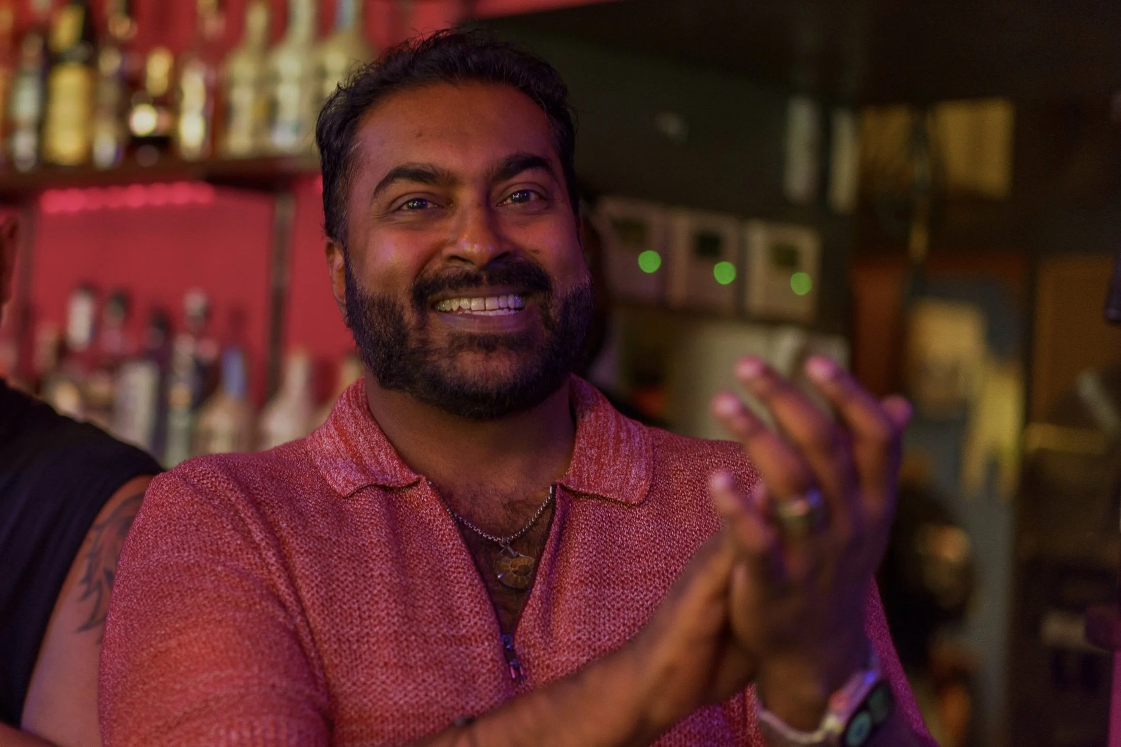 A man with a beard and mustache smiling at a bar, wearing a red patterned shirt, with blurred bottles and bar equipment in the background.