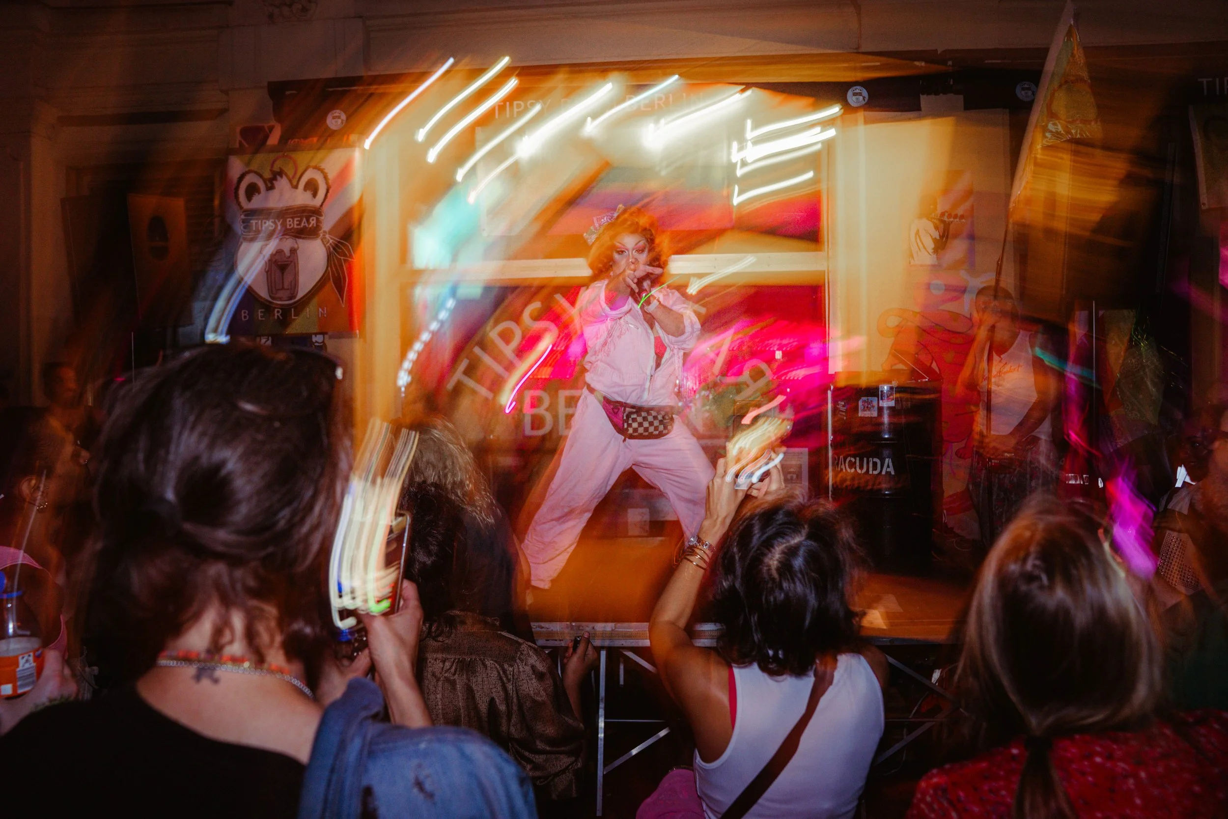 A performer with curly red hair and theatrical makeup gesturing towards the camera on a stage with colorful lights, surrounded by an audience.