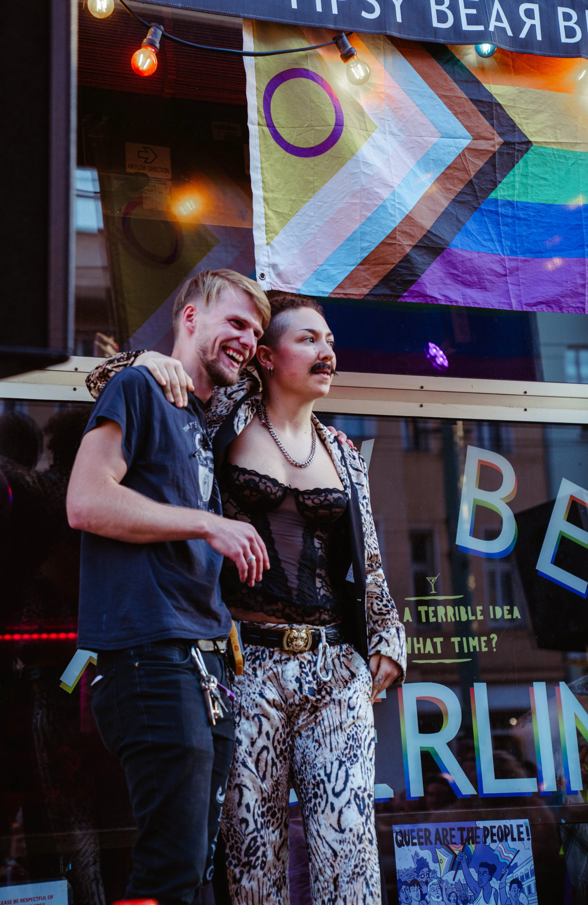 Two people, a man and a woman, standing close together in front of a window display. The man has blond hair and a beard, wearing a navy shirt and pants with keys attached to his belt, smiling and looking to his left. The woman has short dark hair, a 