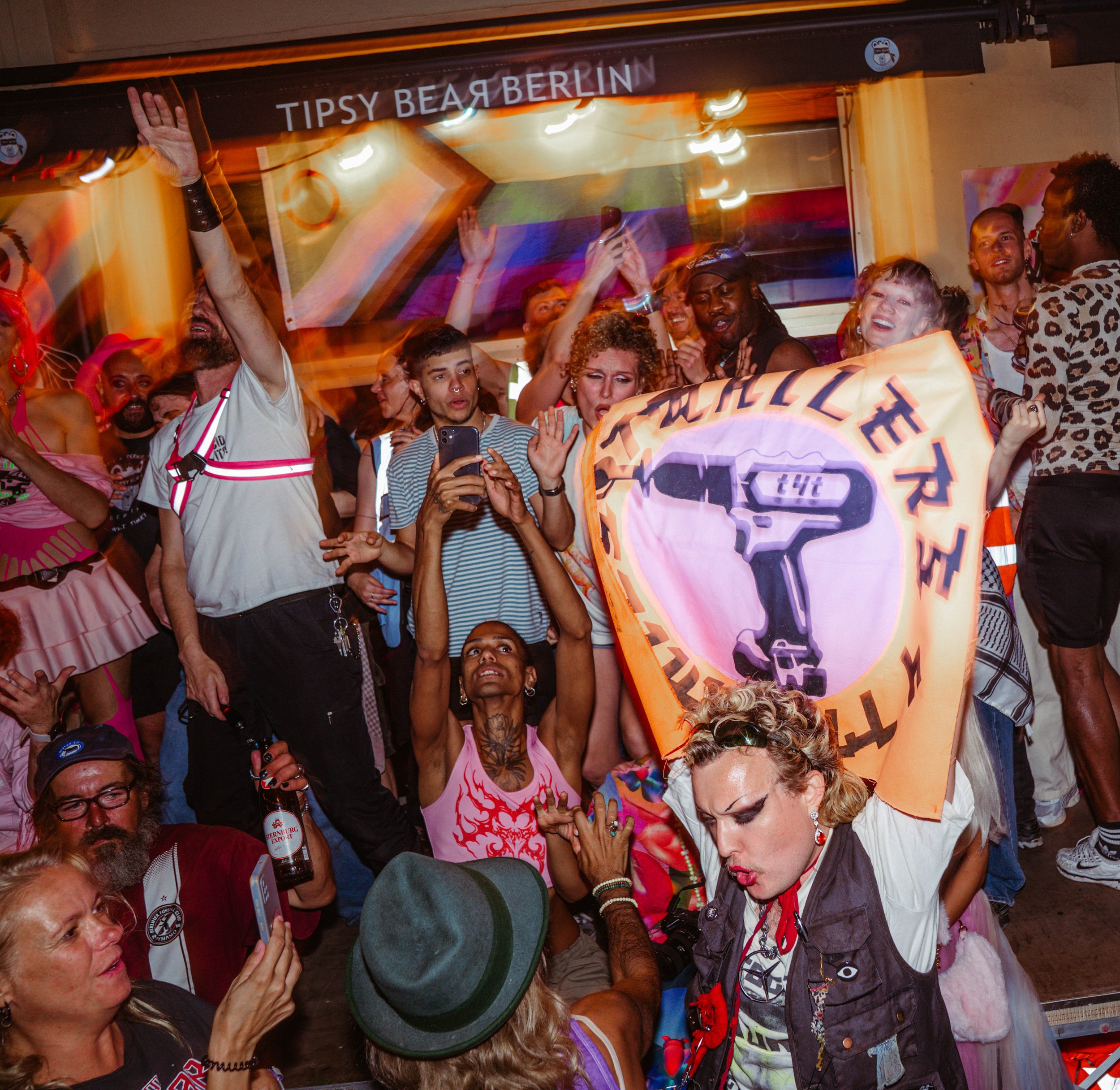 A crowd of diverse people celebrating at a party, some holding cameras and a banner, with bright colorful outfits and expressions of joy.
