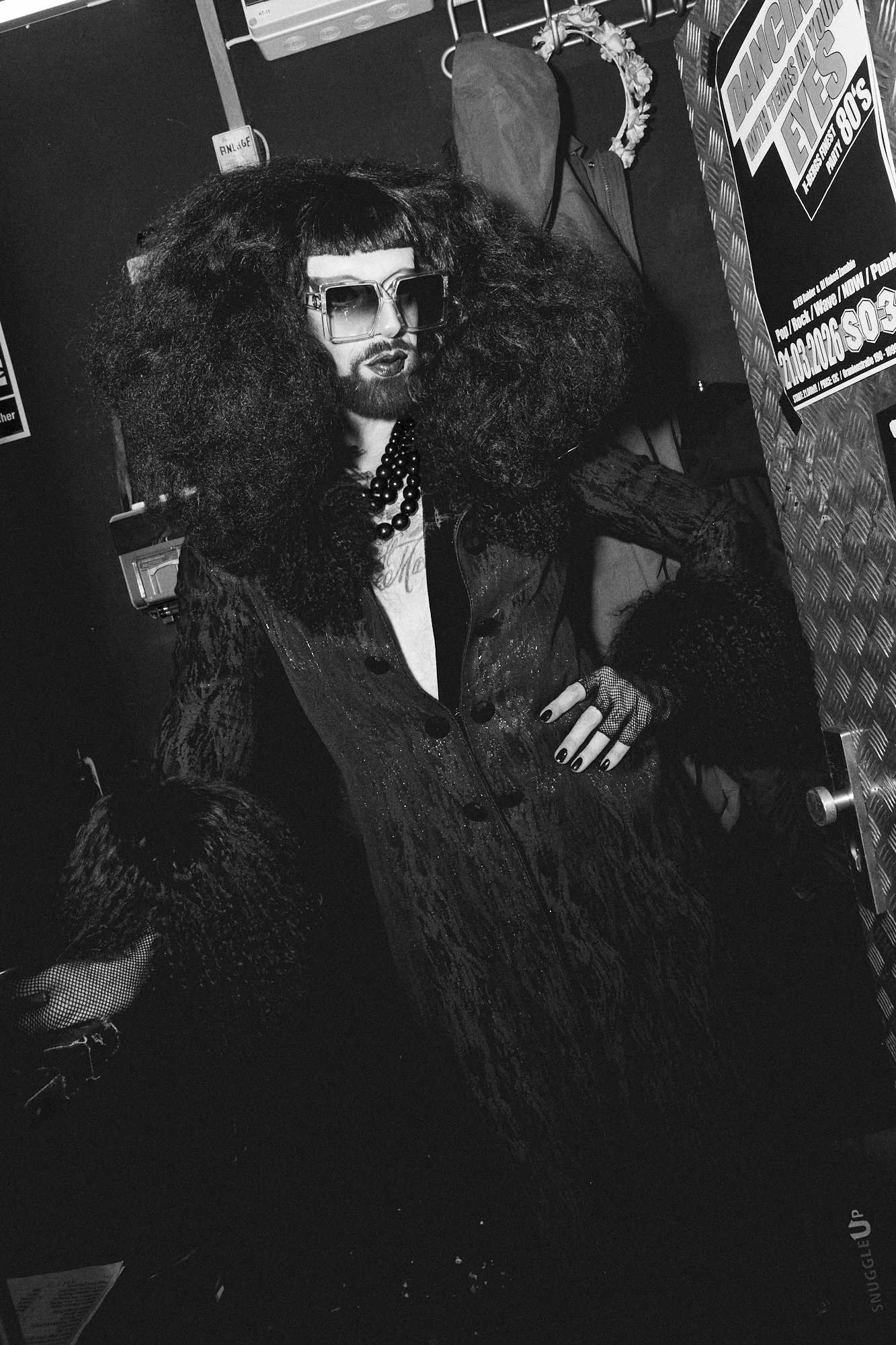 A person with large, curly hair, sunglasses, and a beard, wearing dark clothing with sparkles. They have dark nails and are sitting at a table with promotional signs and items around them in a black and white photo.