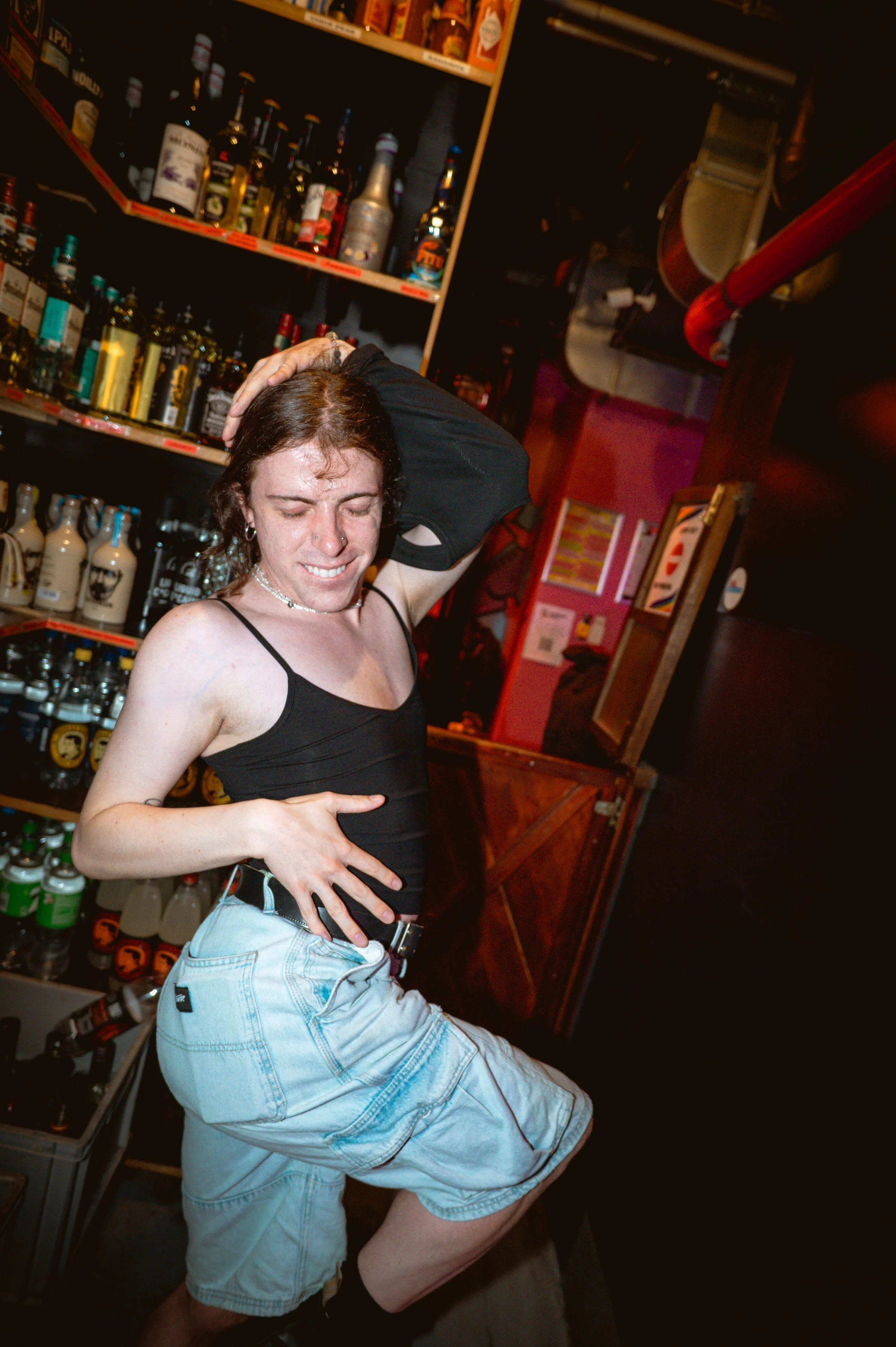 A person with brown hair, wearing a black tank top and light blue shorts, has one hand on their head and the other on their stomach, standing in front of shelves filled with bottles of alcohol in a bar or liquor store.