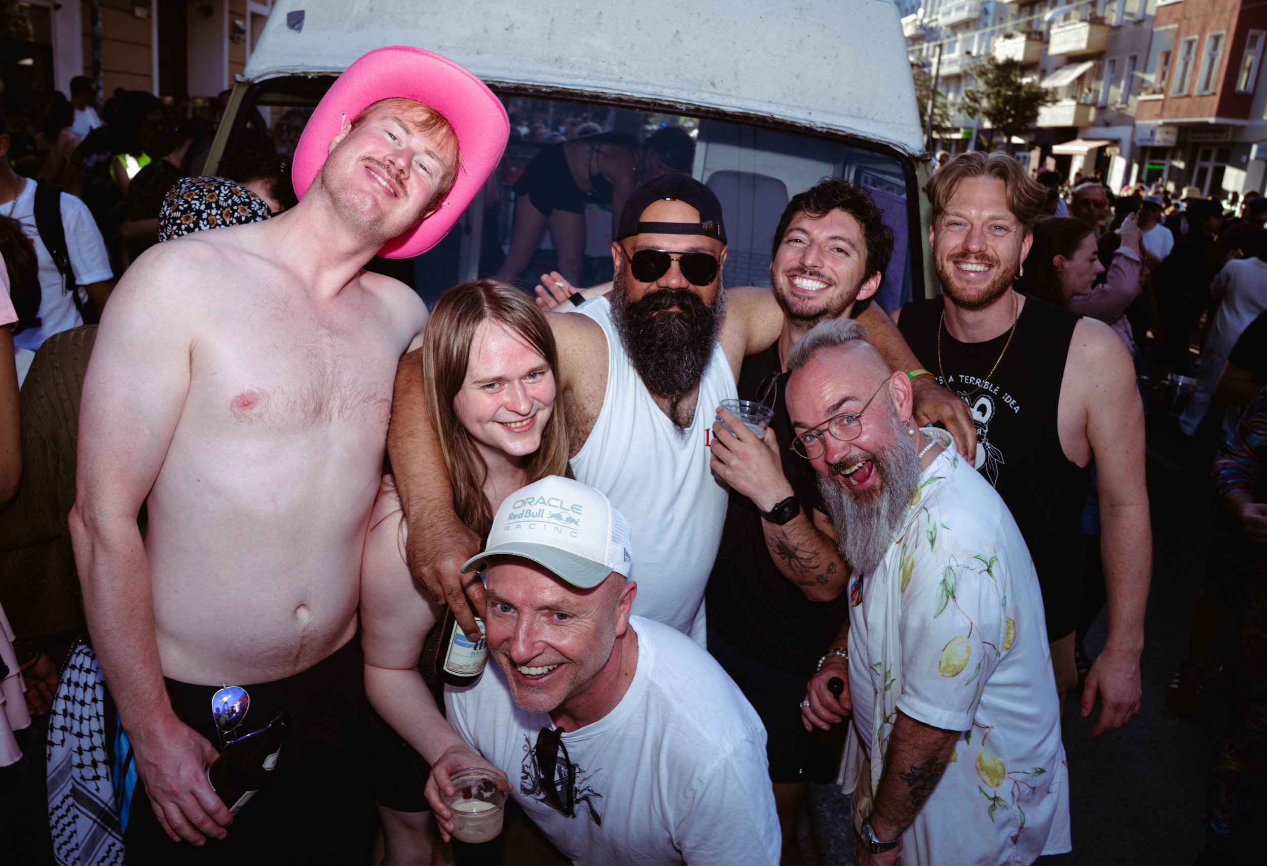 Group of friends at a lively outdoor event, smiling and engaging with each other, some holding drinks, with a crowded city street backdrop.