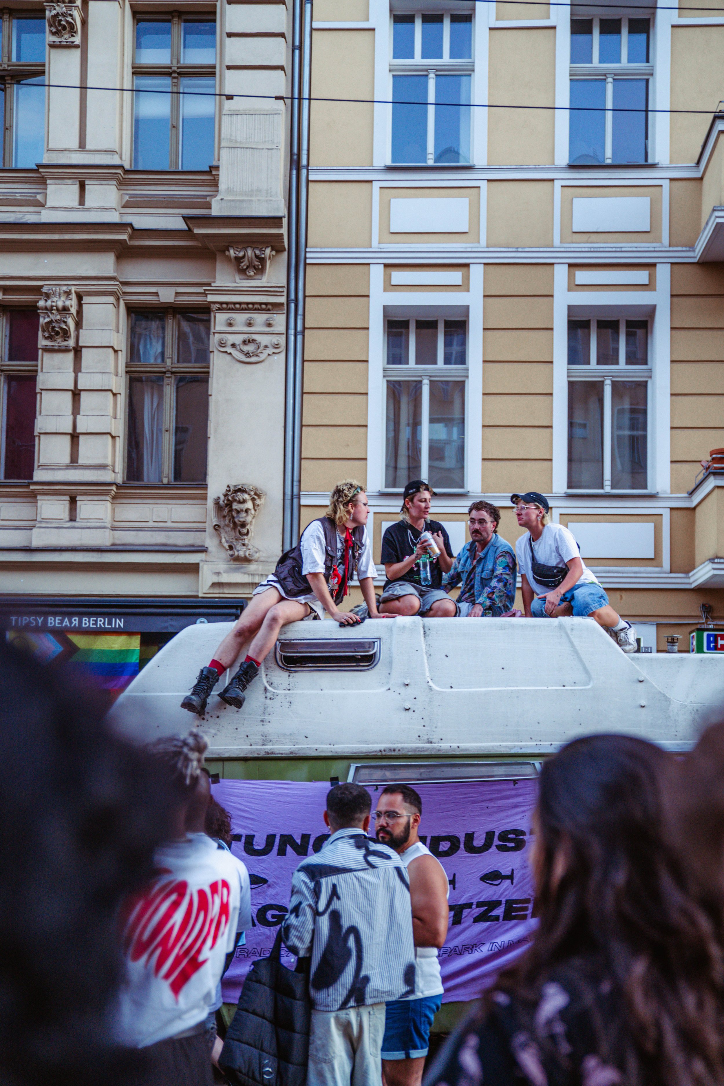 A group of young people sitting and standing on top of a vehicle during a parade, with onlookers in the foreground and multi-story buildings in the background.