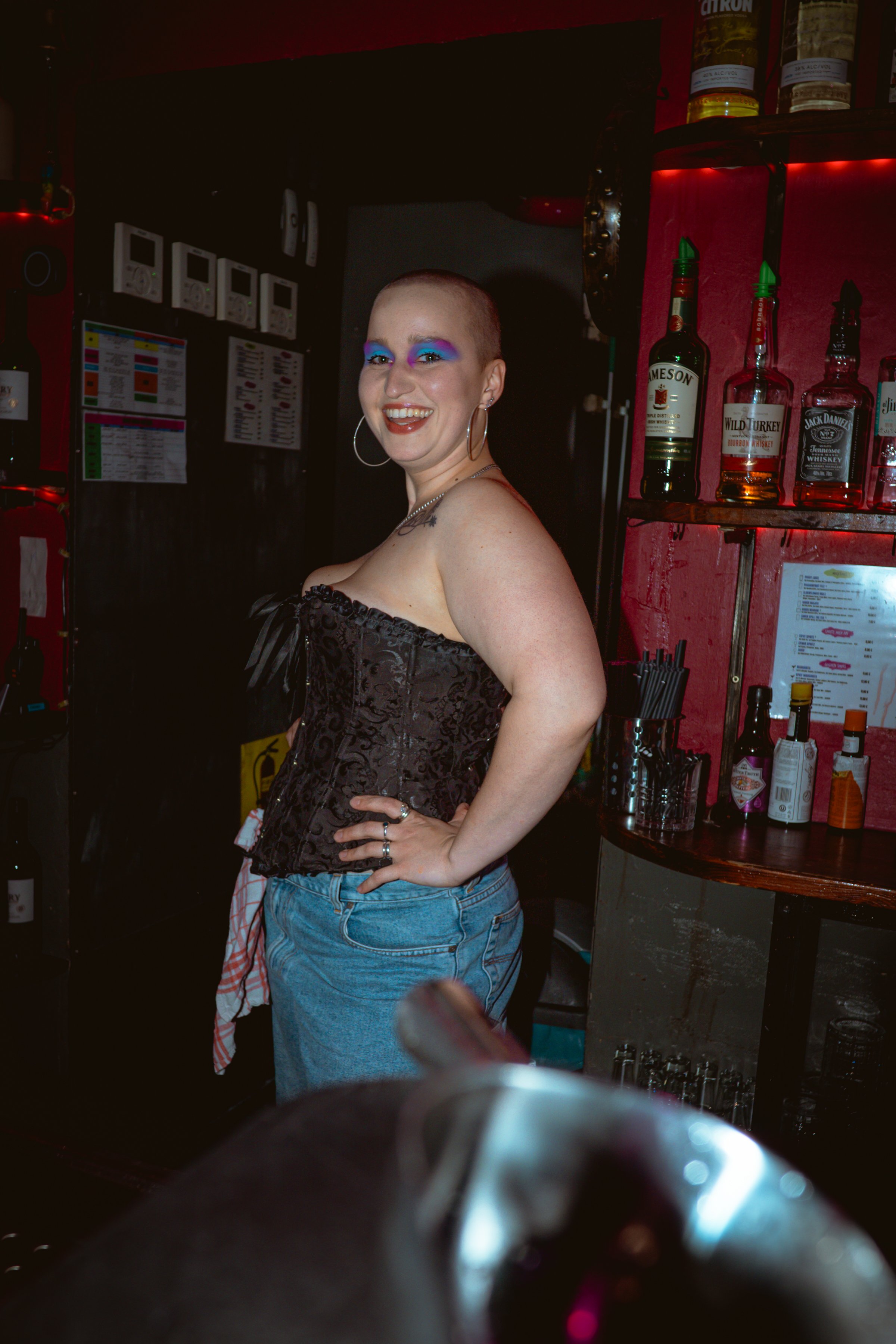 A woman with a shaved head and colorful eye makeup smiling in a bar setting, with bottles of alcohol behind her and a red wall.