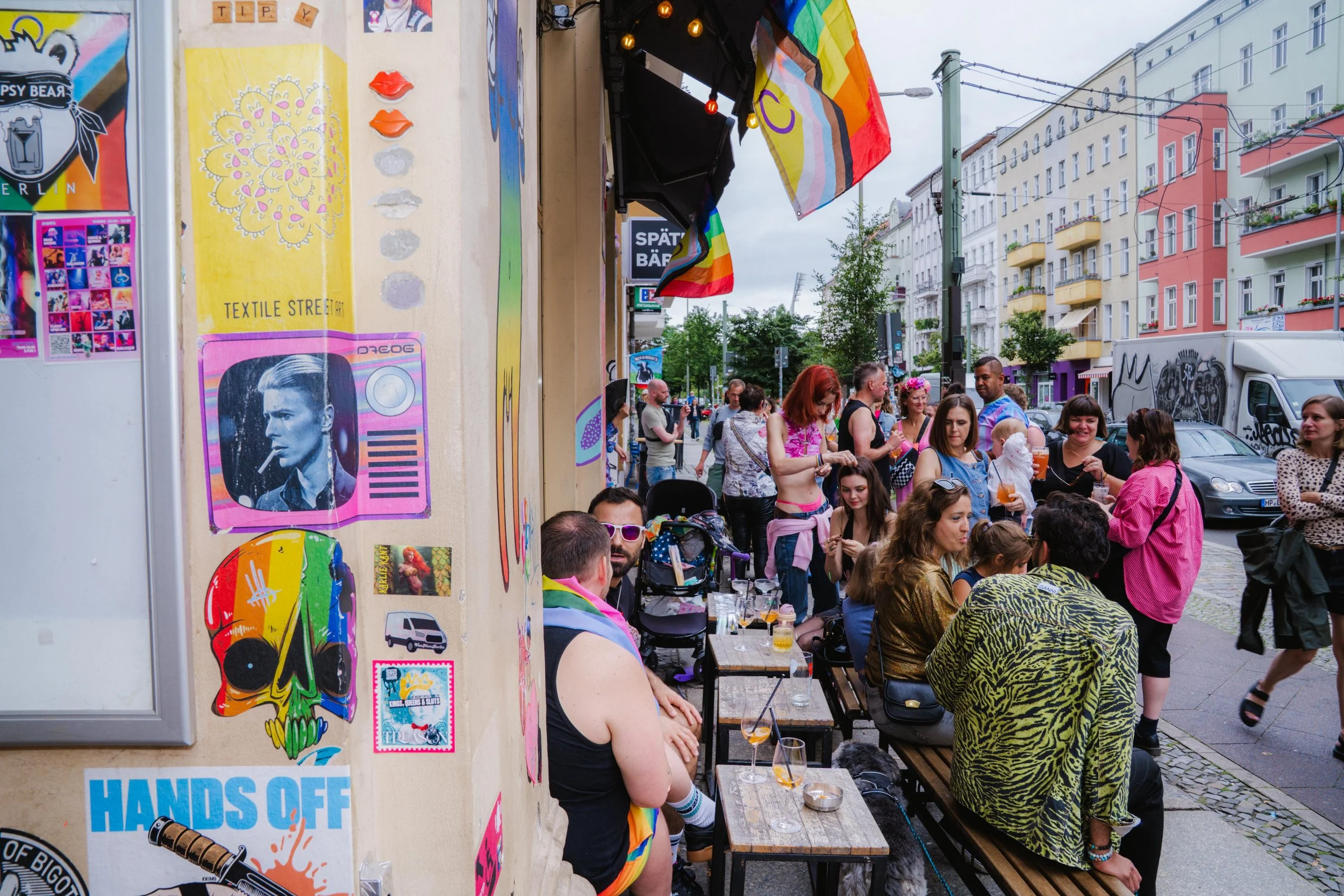 A busy outdoor street scene with a group of people socializing at a table and standing around. The scene is decorated with rainbow flags and various colorful artwork on a wall, including a poster of a man with a cigarette. There are multiple modern a