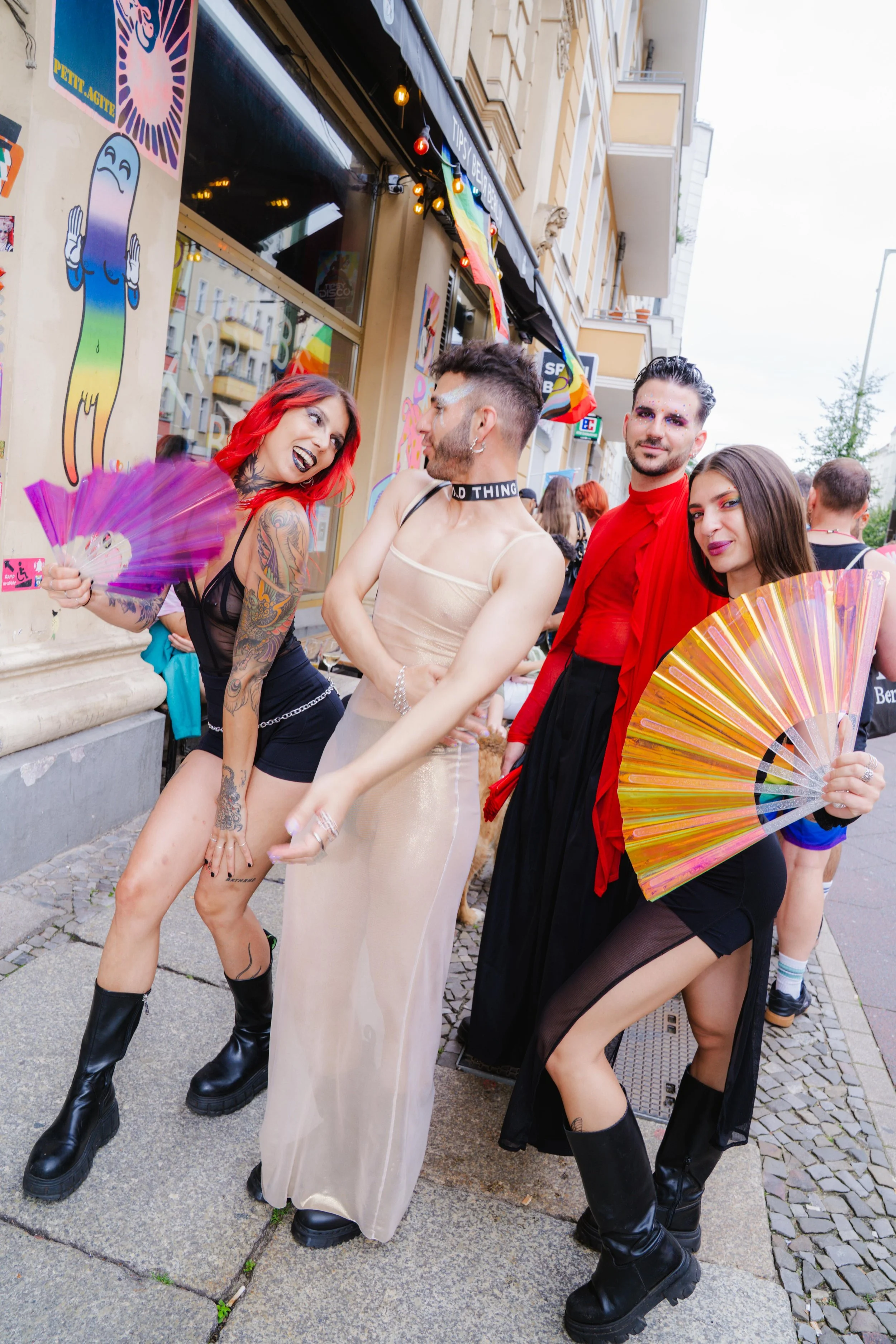 Five people with bold and colorful fashion styles, including tattoos, makeup, and accessories, standing on a city street during an outdoor event or parade. They are smiling, engaging with each other, and holding rainbow-colored fans. The scene includ
