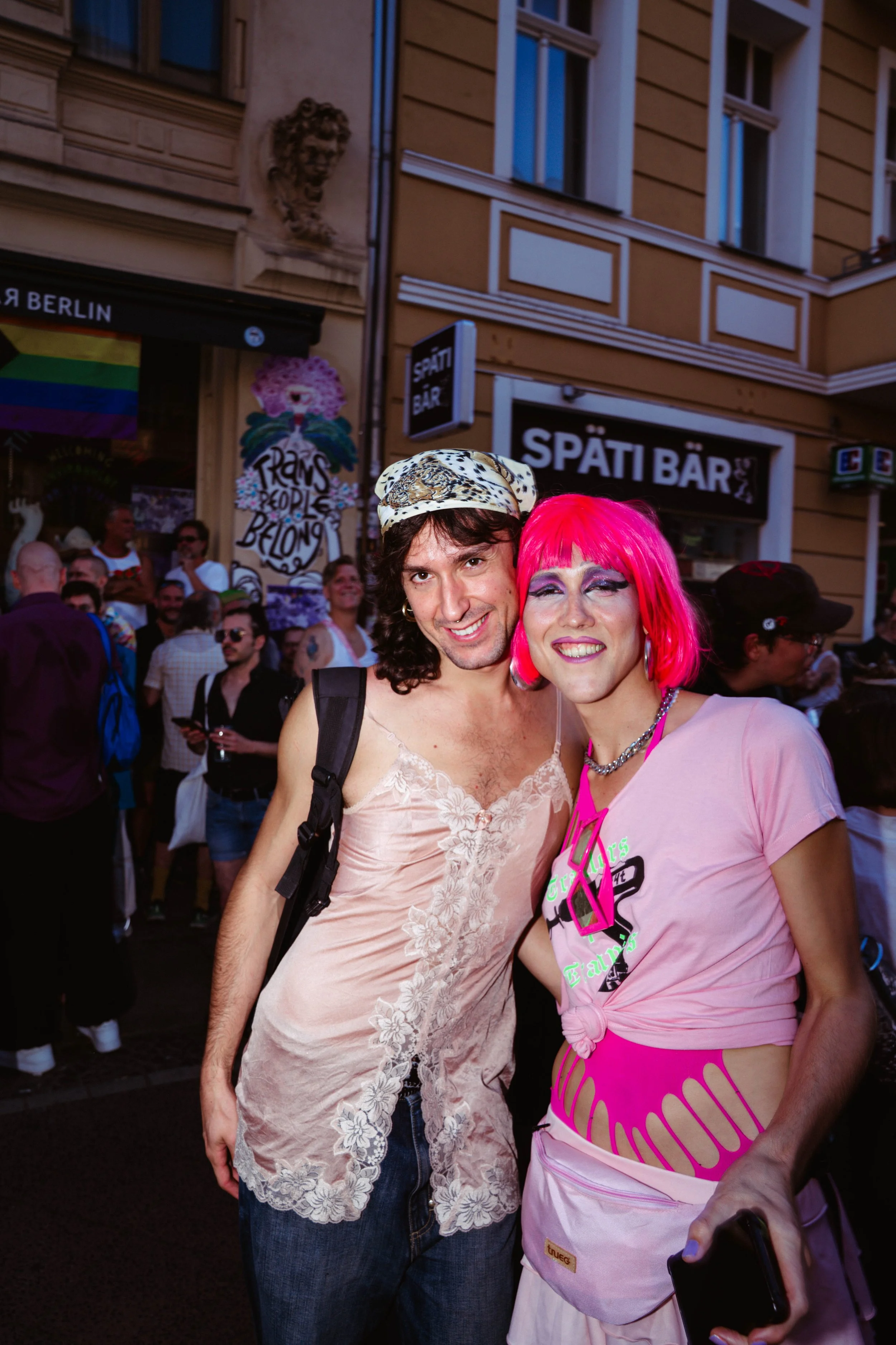 Two people dressed in vibrant, eye-catching clothing and wigs standing close together and smiling at a busy outdoor event in an urban area with many other attendees, shops, and signage in the background.