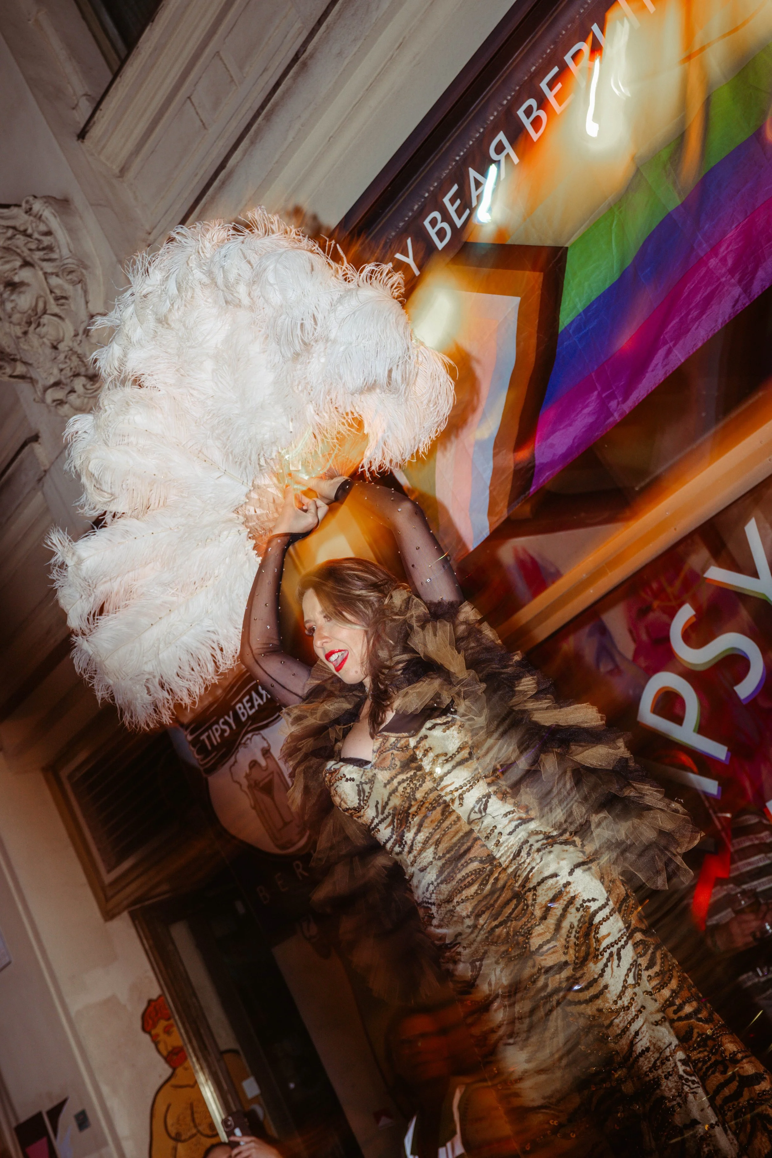 A person with dark hair and red lipstick in a glittery gold and black dress. She is smiling and posing with a woman dressed as a large white feathered bird in a venue with a colorful rainbow banner and room decor.