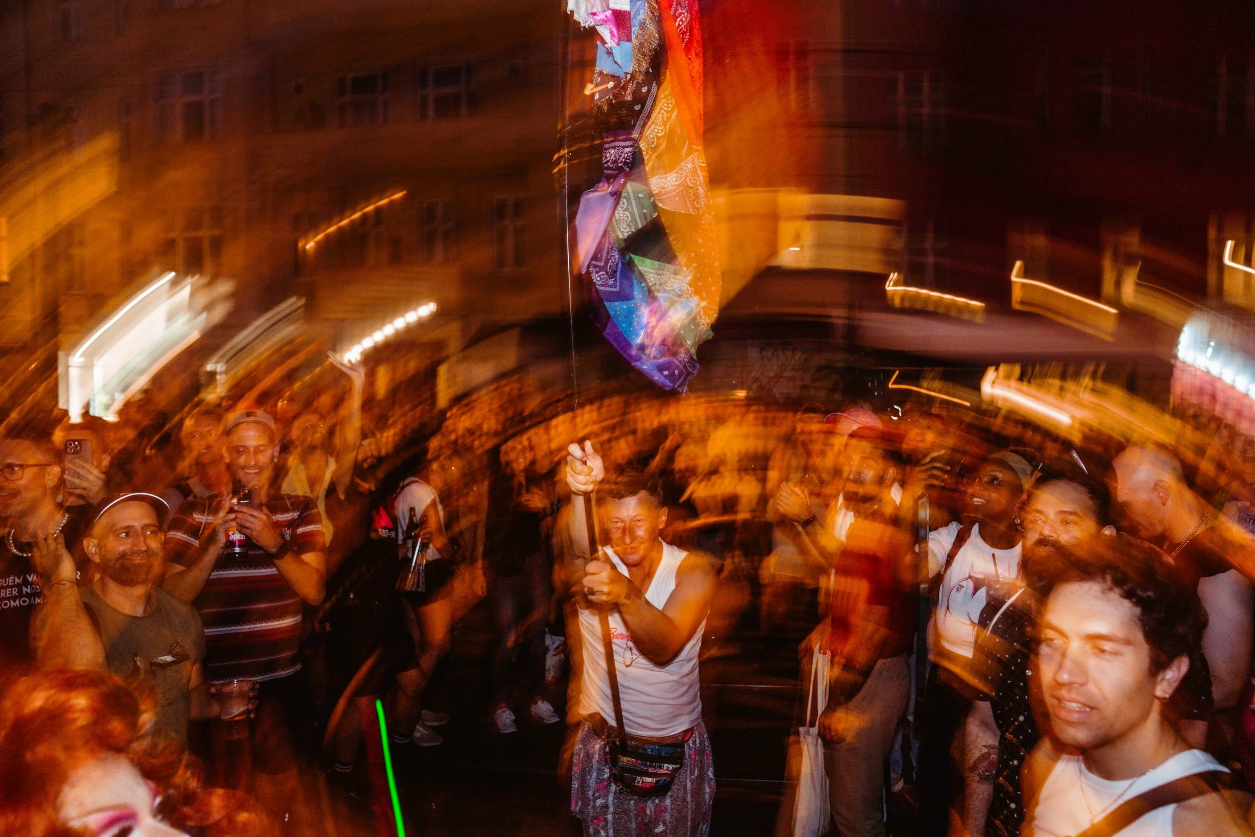 A lively crowd of people at a nighttime event, with some holding drinks and others taking photos. The scene features motion blur, suggesting movement and excitement, with a colorful fabric or banner hanging in the background.