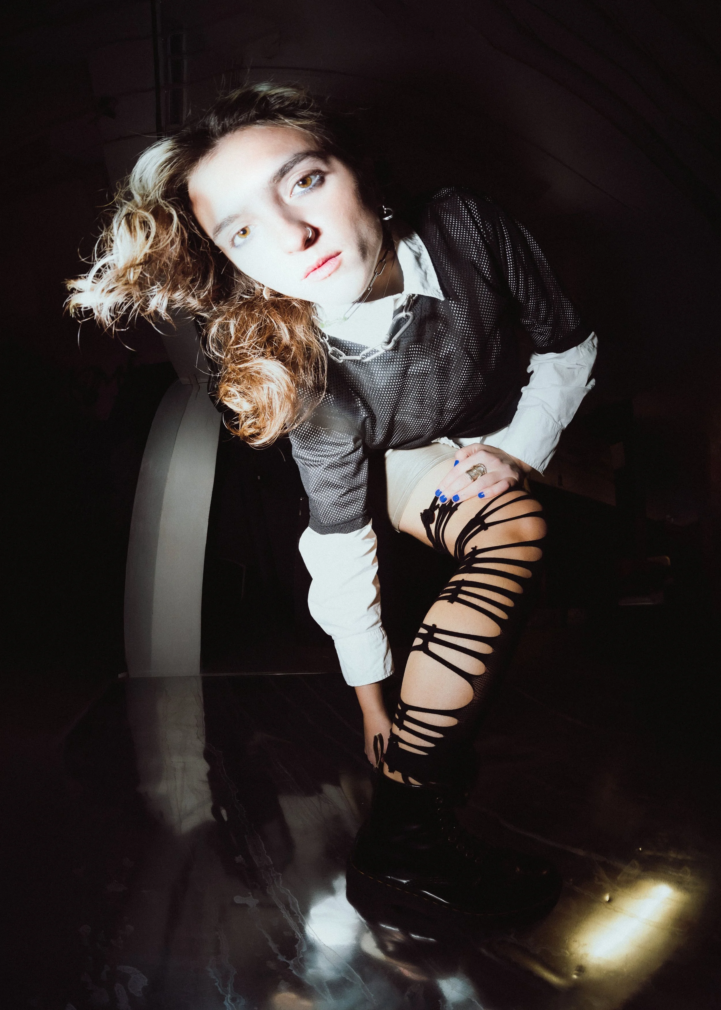 A young person with a nose ring and wavy hair, dressed in a black mesh top over a white shirt, beige shorts, and black lace-up boots, posing in a dark room with an intense gaze.