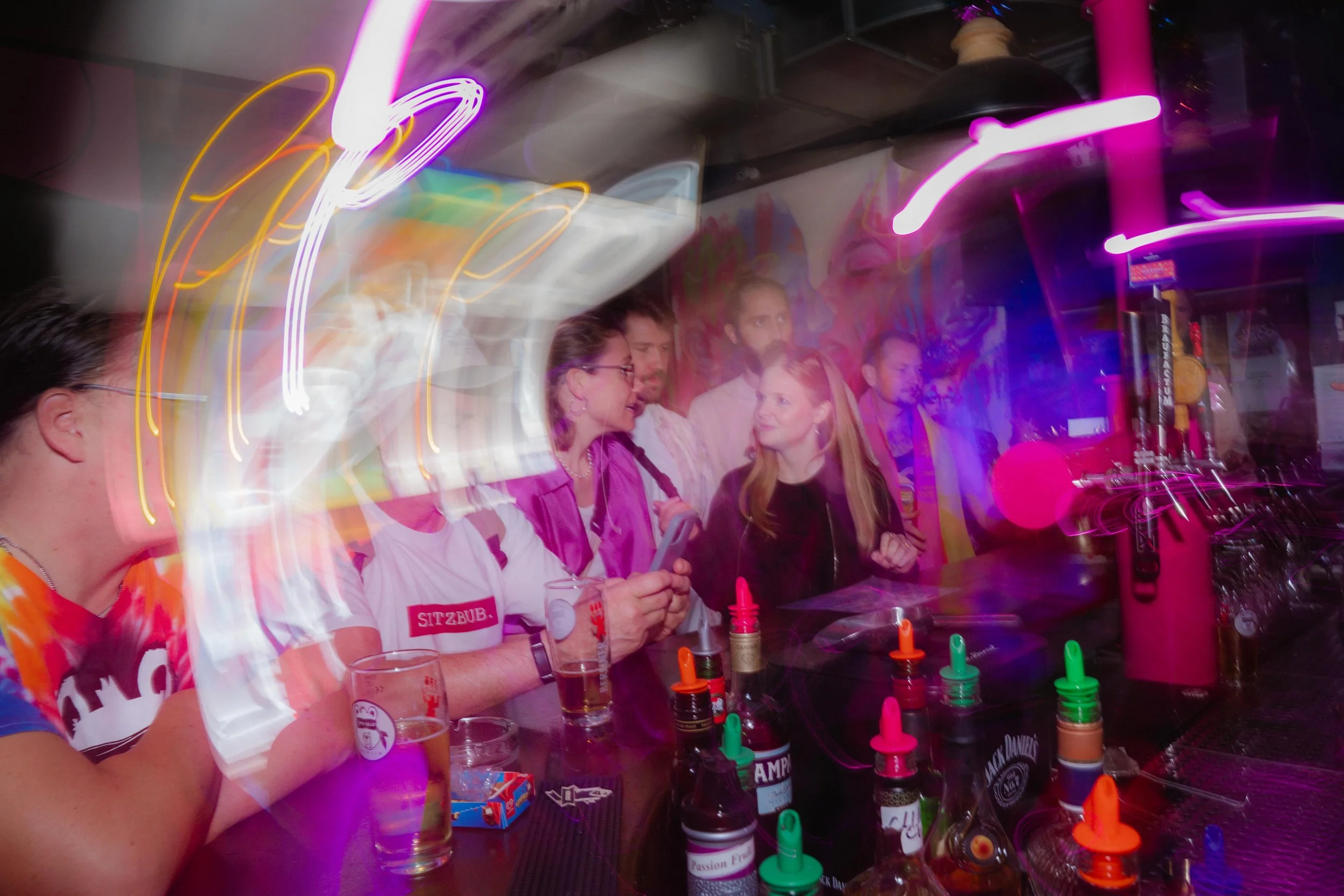 A group of people standing at a bar counter with neon and colorful lights in the background, engaged in conversation. Various bottles and condiment bottles are on the counter.