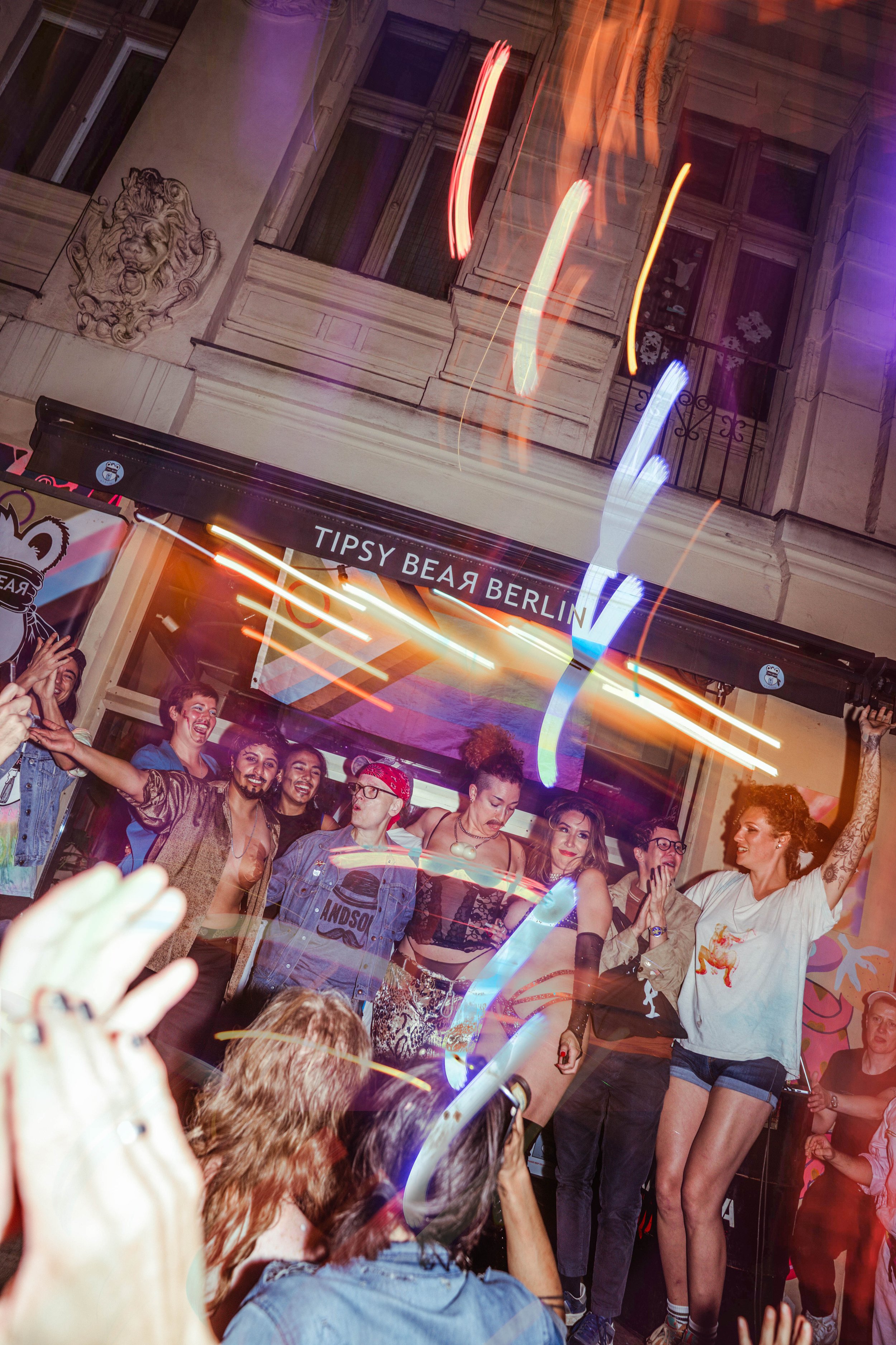 Crowd of people at a lively party or club with colorful lights, smiling and dancing, with a building facade in the background.