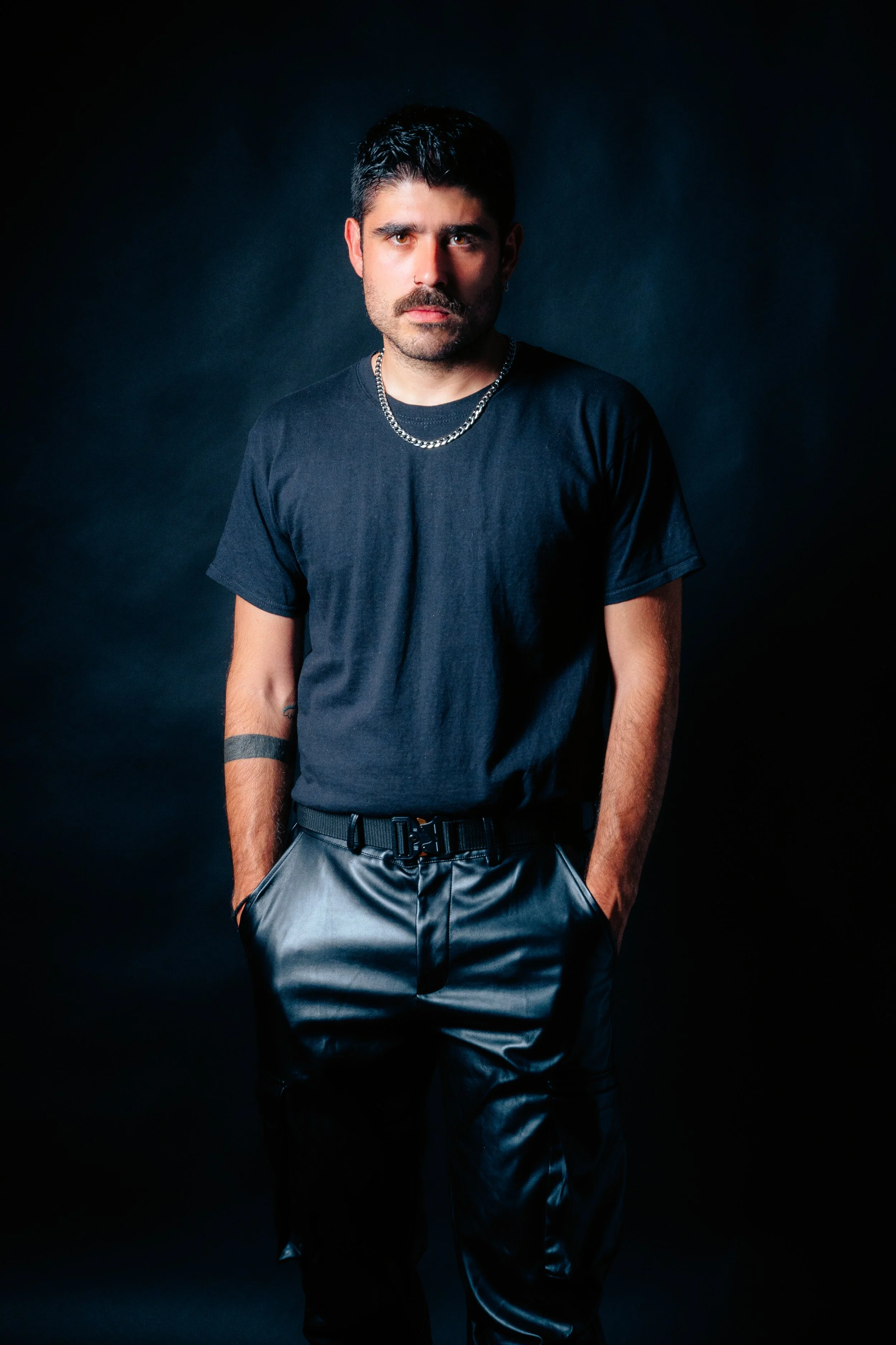 Portrait of a young man with dark hair and a mustache, wearing a black t-shirt and shiny black pants, standing against a dark background with hands in pockets, looking directly at the camera.