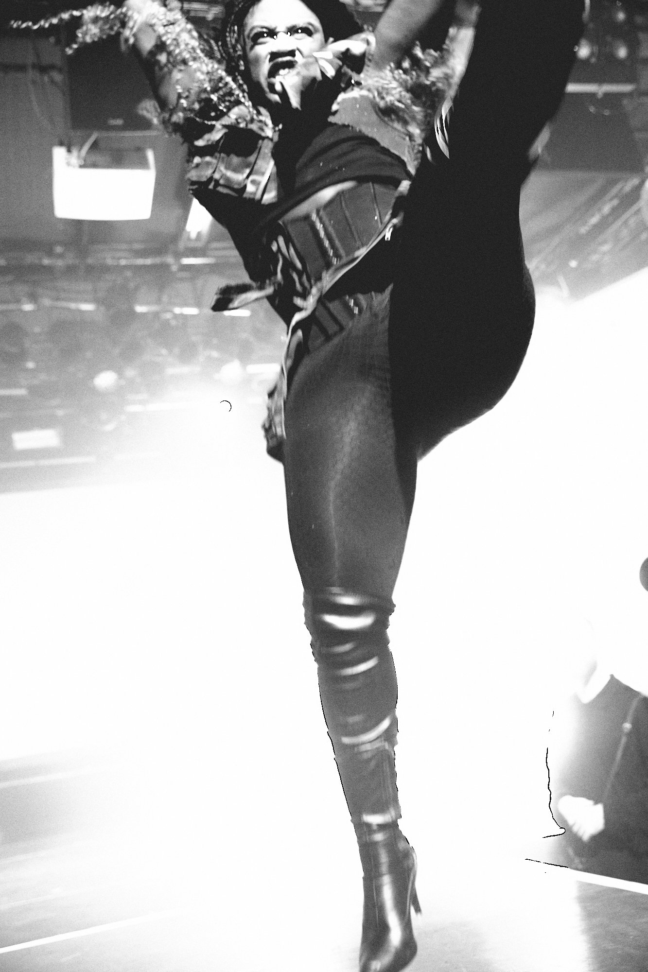 A performer dressed in a patterned jacket, black top, and tight shiny pants, performing a dance move on stage, captured in black and white.