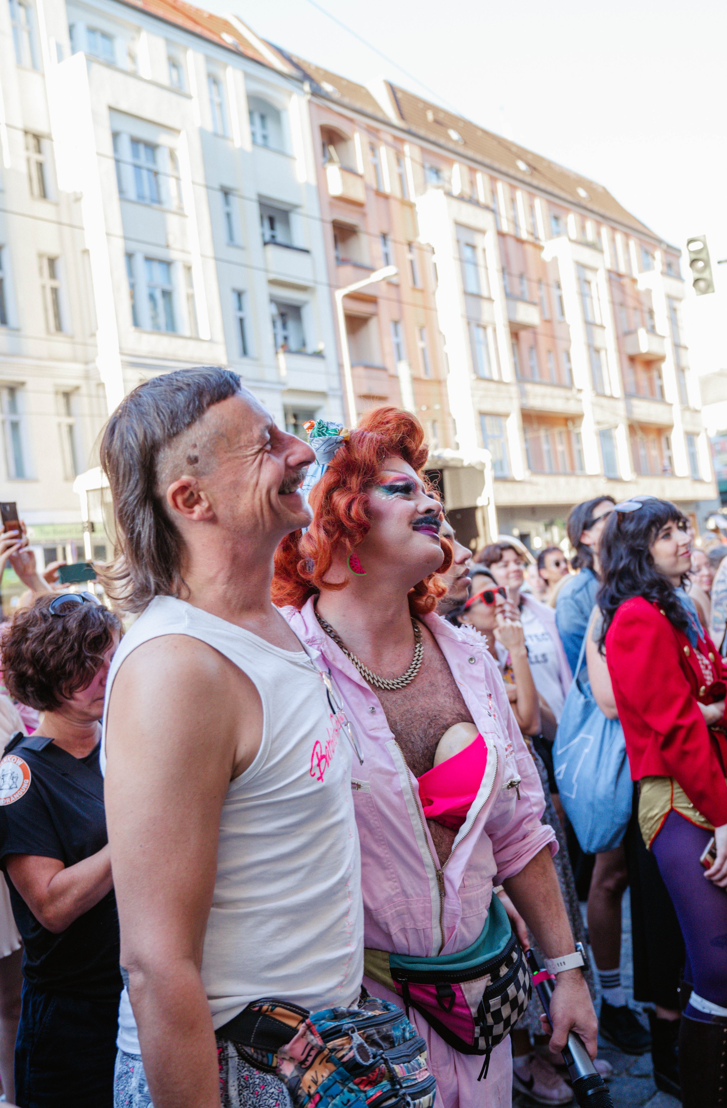 Crowd of people at an outdoor event, some dressed in colorful costumes, with buildings and a traffic light in the background.