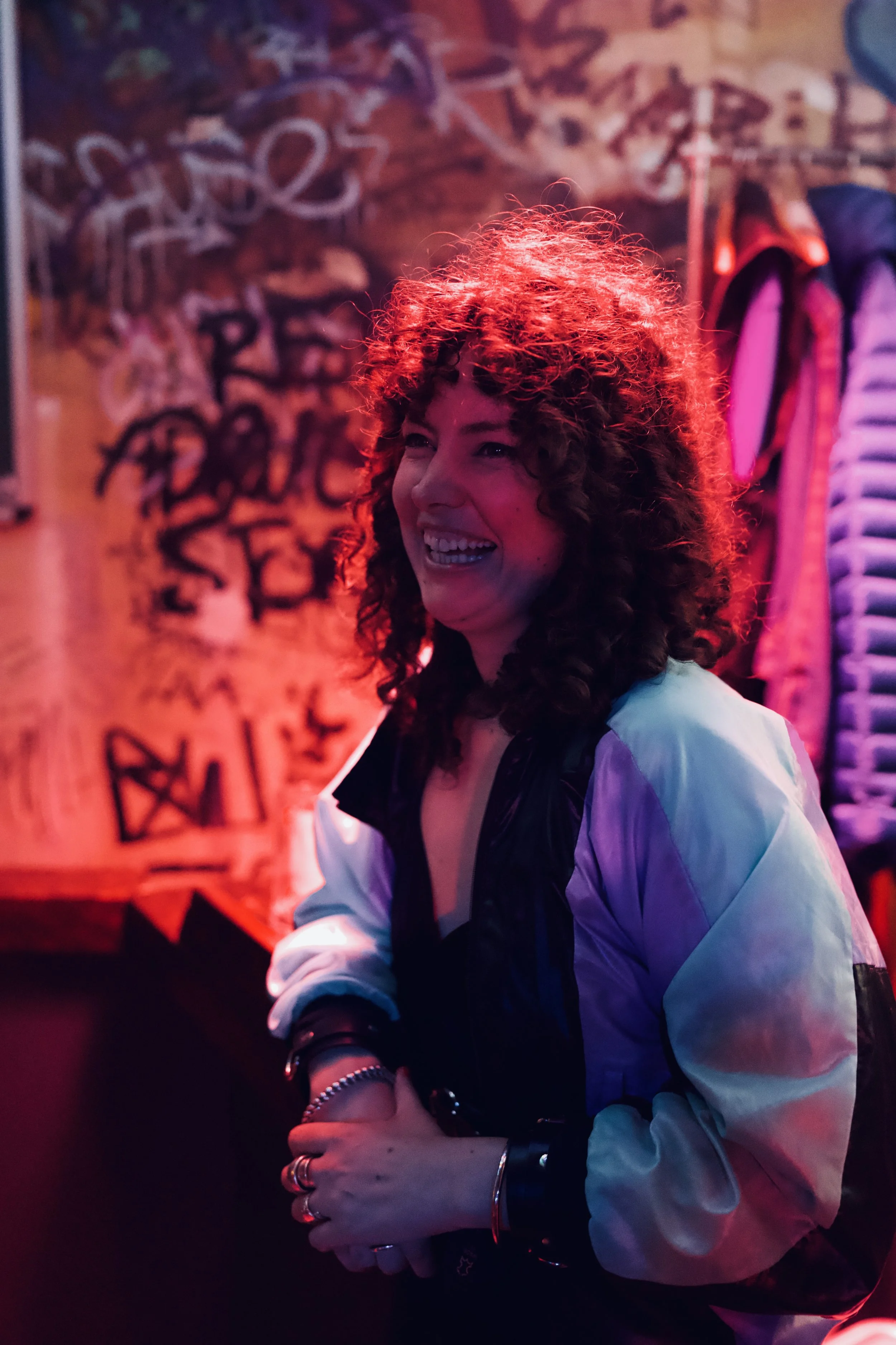 A woman with curly hair, smiling, in a dimly lit room with red and purple lighting, graffiti on the wall behind her, and various clothing items hanging.