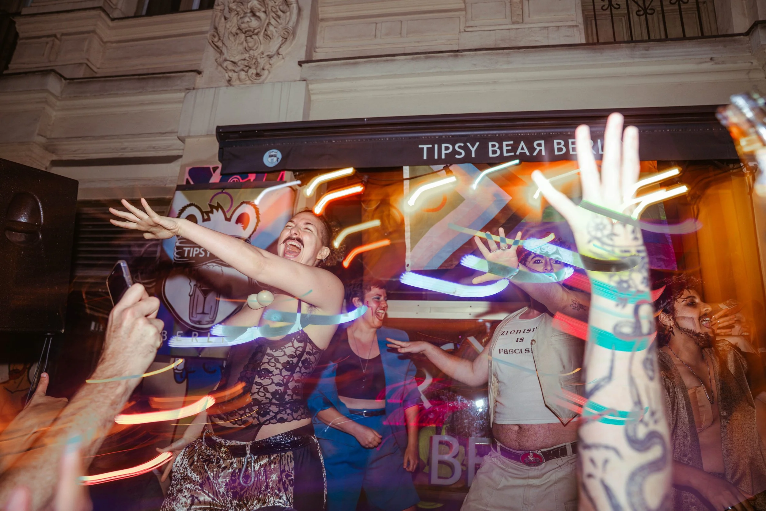 A group of people celebrating inside a bar with colorful lights, some with their hands raised, smiling and dancing.