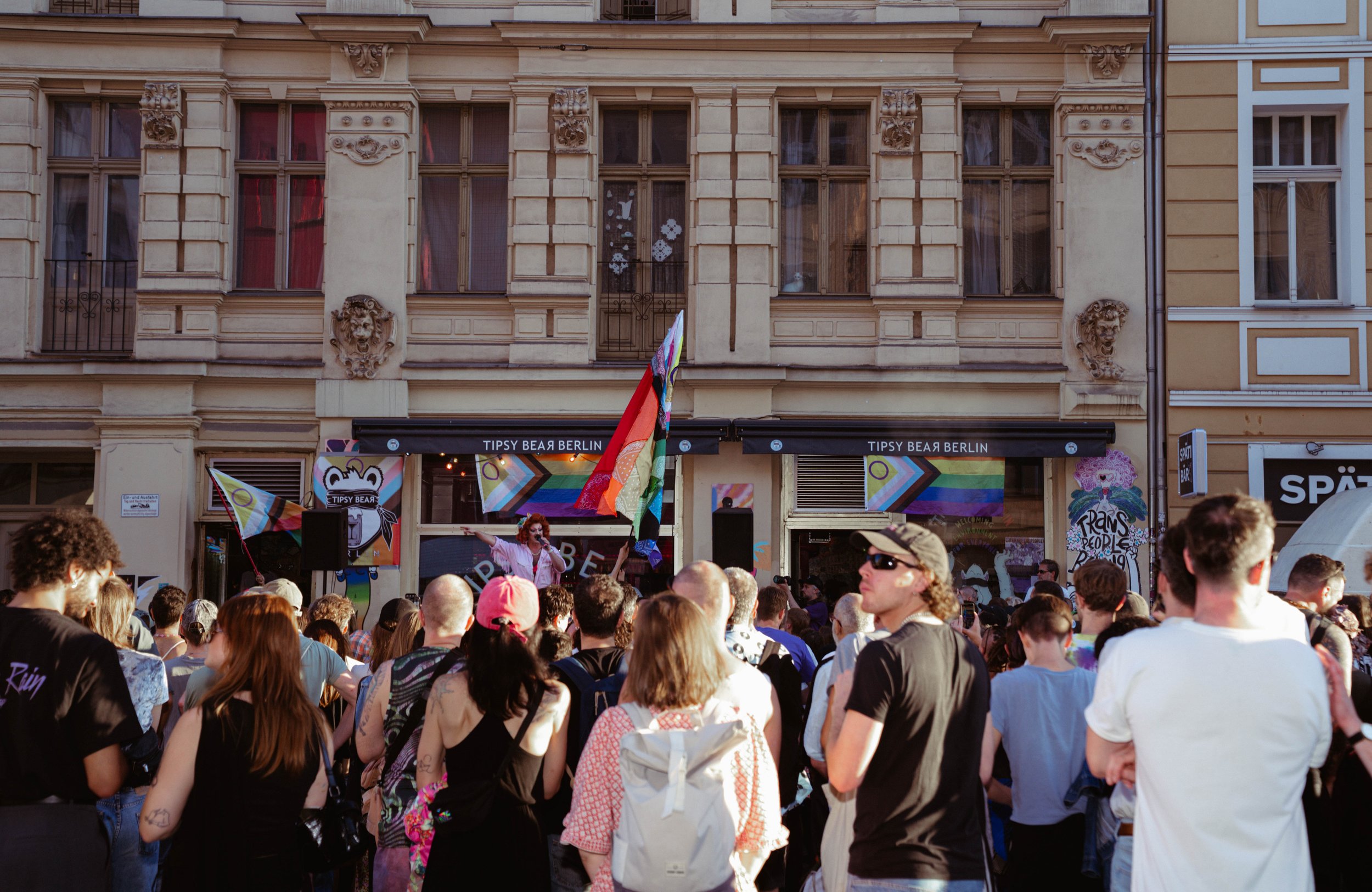 Crowd gathered in front of a building with celebration flags, a person speaking or singing into a microphone, and rainbow pride flags.
