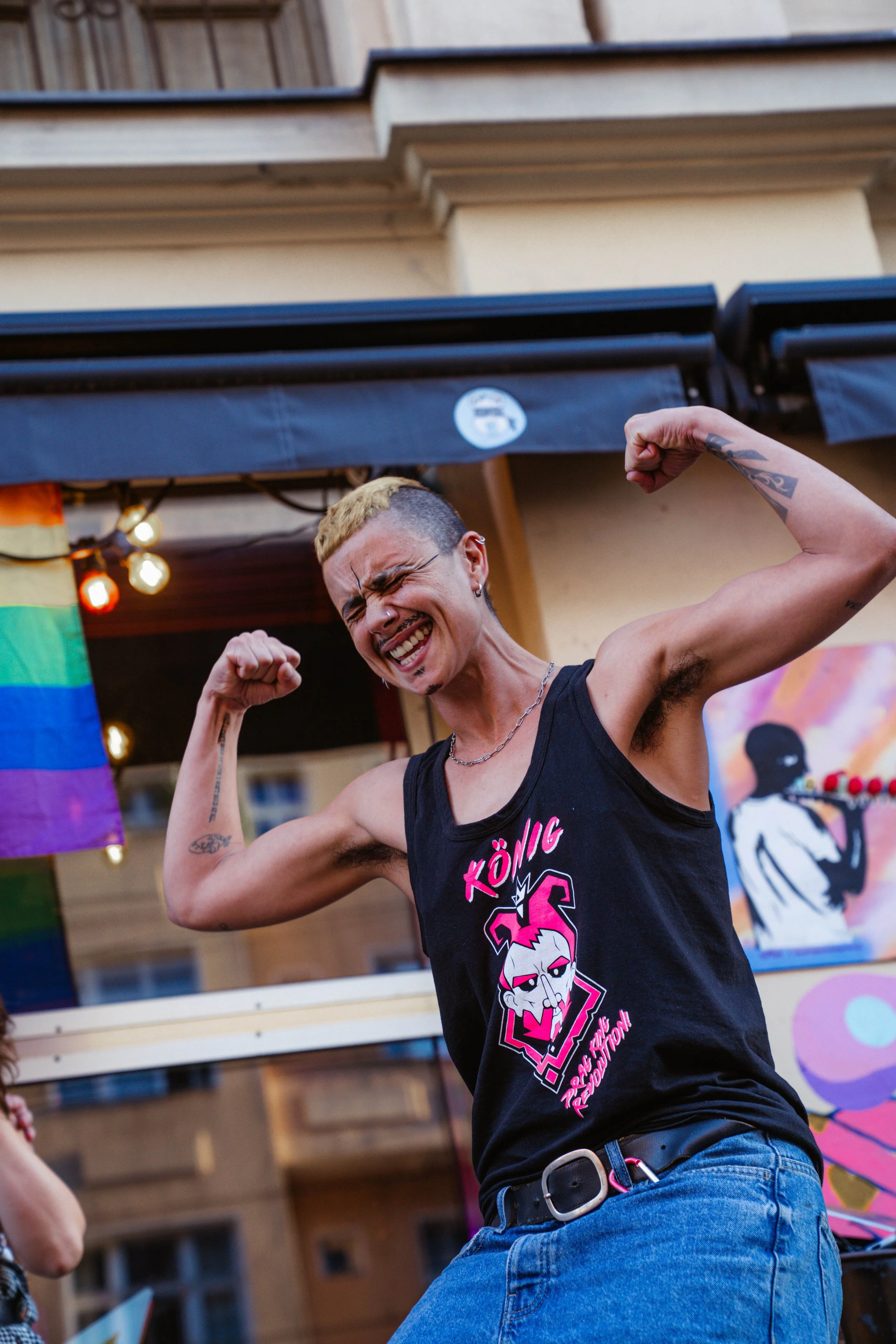Person with short hair, tattoos, and jewelry, flexing muscles and smiling. Wearing a black tank top with graphics and blue jeans, inside a building with colorful decorations and flags.