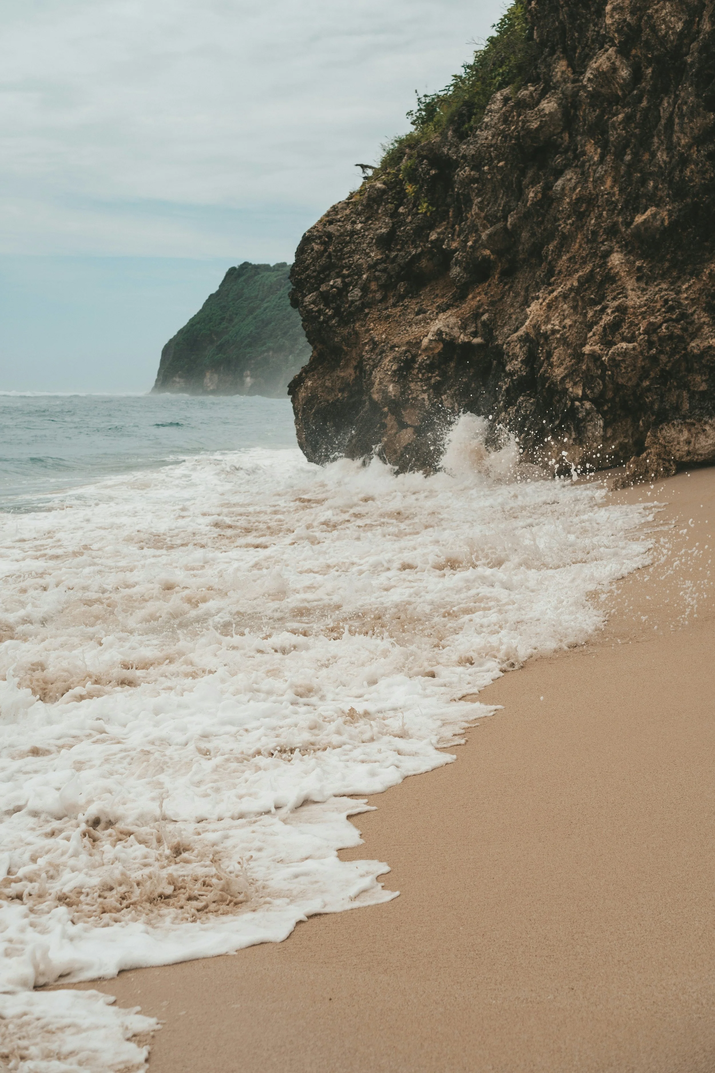 A sandy beach with waves crashing against large dark brown cliffs, some green vegetation on the cliffs, and a cloudy sky overhead.