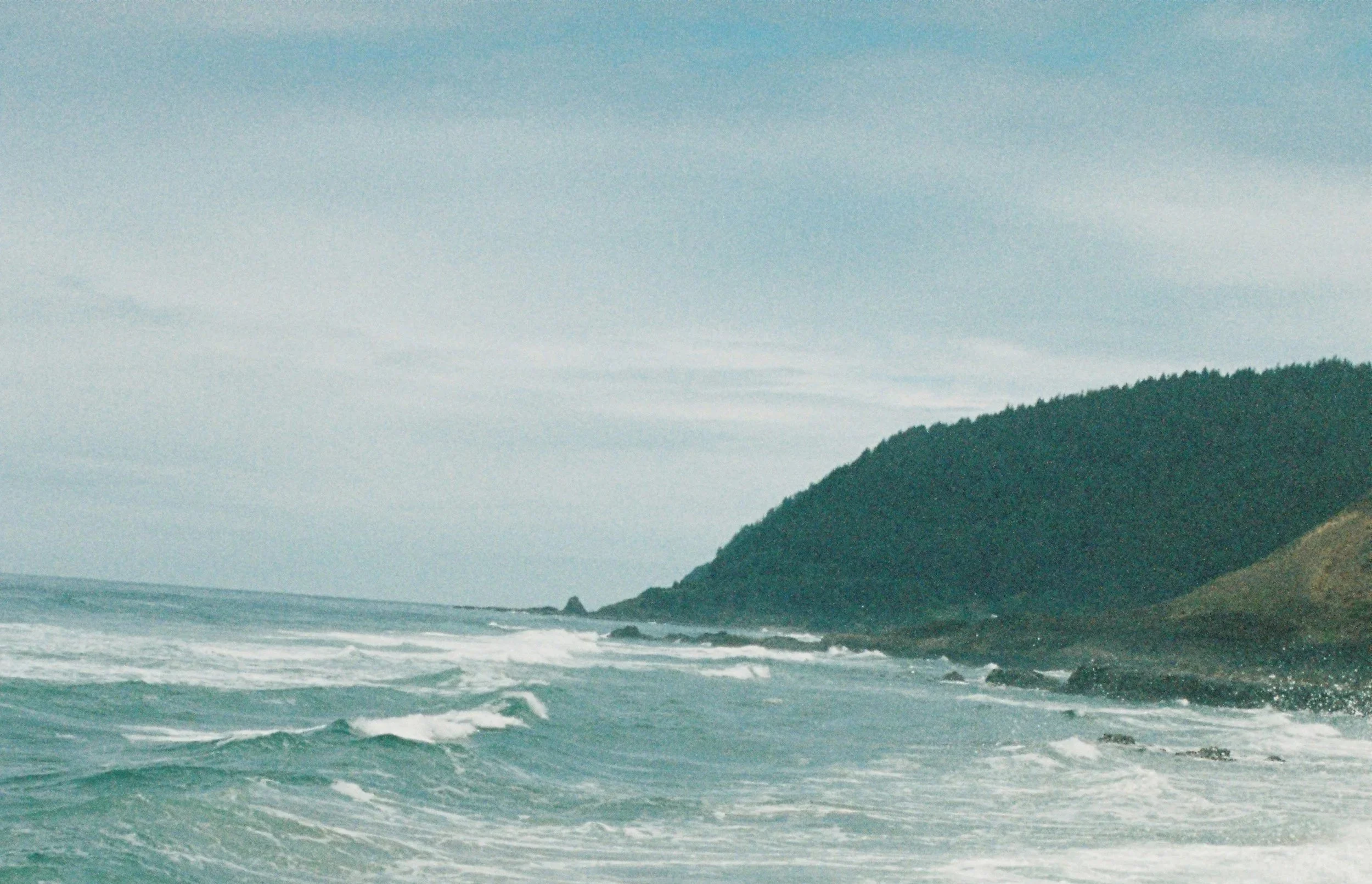 A view of the ocean with waves crashing against the shoreline and forested hills in the background under a cloudy sky.