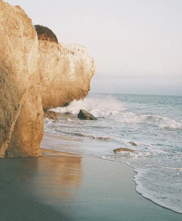Cliffside view of a beach with waves crashing against rocks and sandy shore at sunset or sunrise.