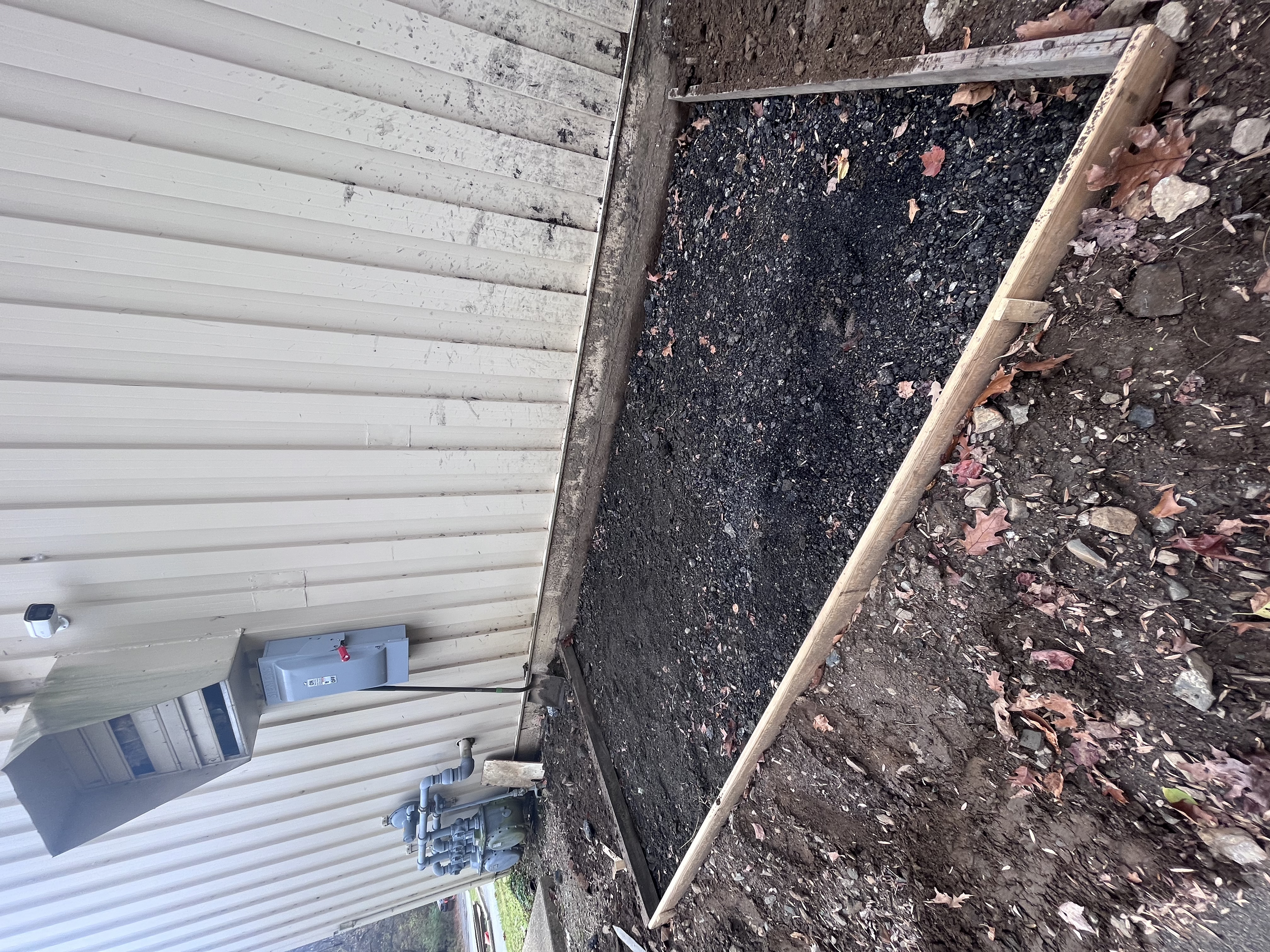 Construction area next to a beige metal building with pipes and utility meters on the wall, soil and gravel in the foreground, and a black pipe running along the ground.