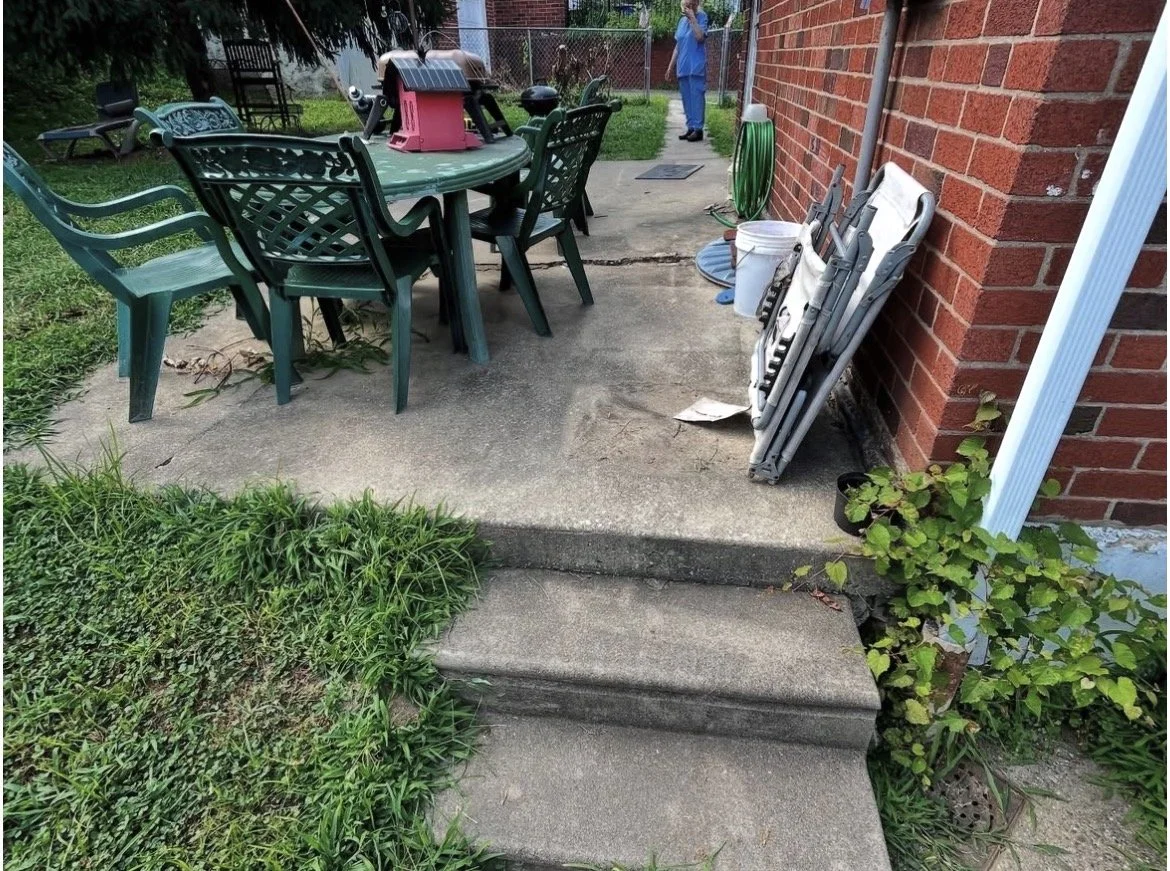 Backyard patio with green plastic table and chairs, folded white patio furniture leaning against a brick wall, a white bucket, garden hose, small plant, and person in blue scrubs in the background