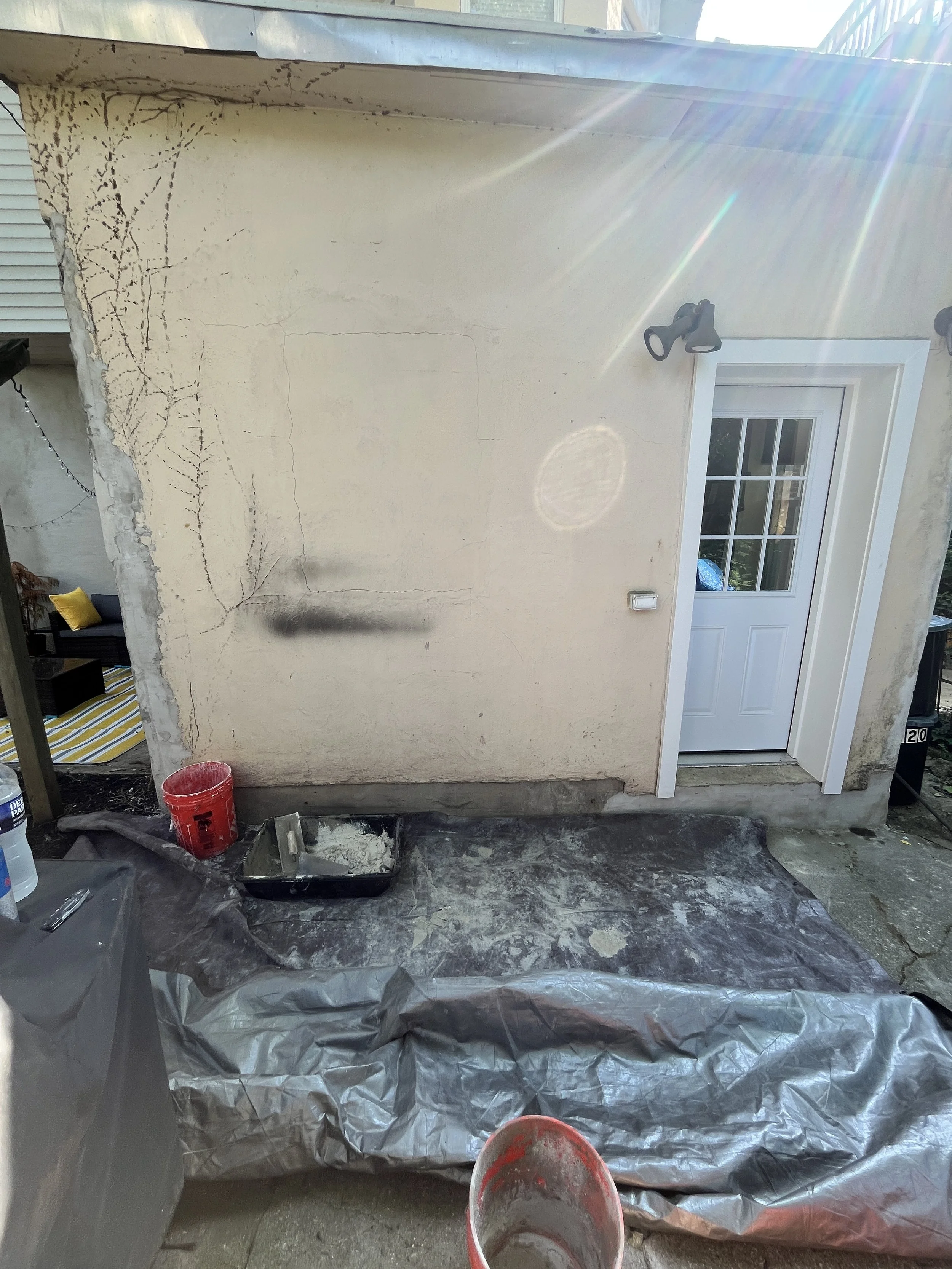Exterior wall of a house being repaired, with visible cracks and patching. Construction tools and supplies, including a red bucket, black tray, and gray plastic sheeting, are on the ground in front of the wall. Sunlight beams across the scene.