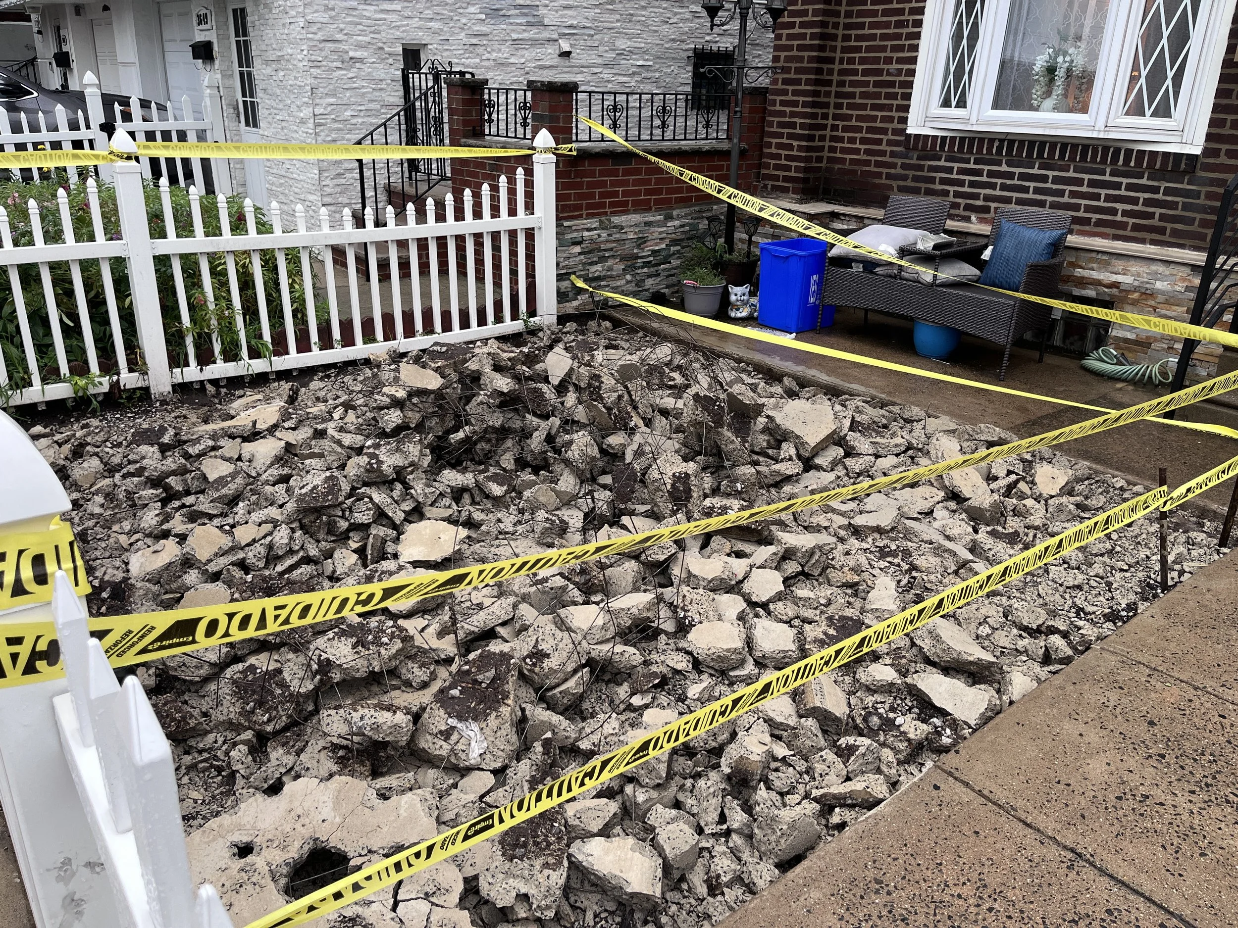 A front yard with a concrete sidewalk, white picket fence, and brick house with a large window. The yard is cordoned off with yellow caution tape, and there is a large pile of broken concrete and debris on the ground. A blue recycling bin, a potted p