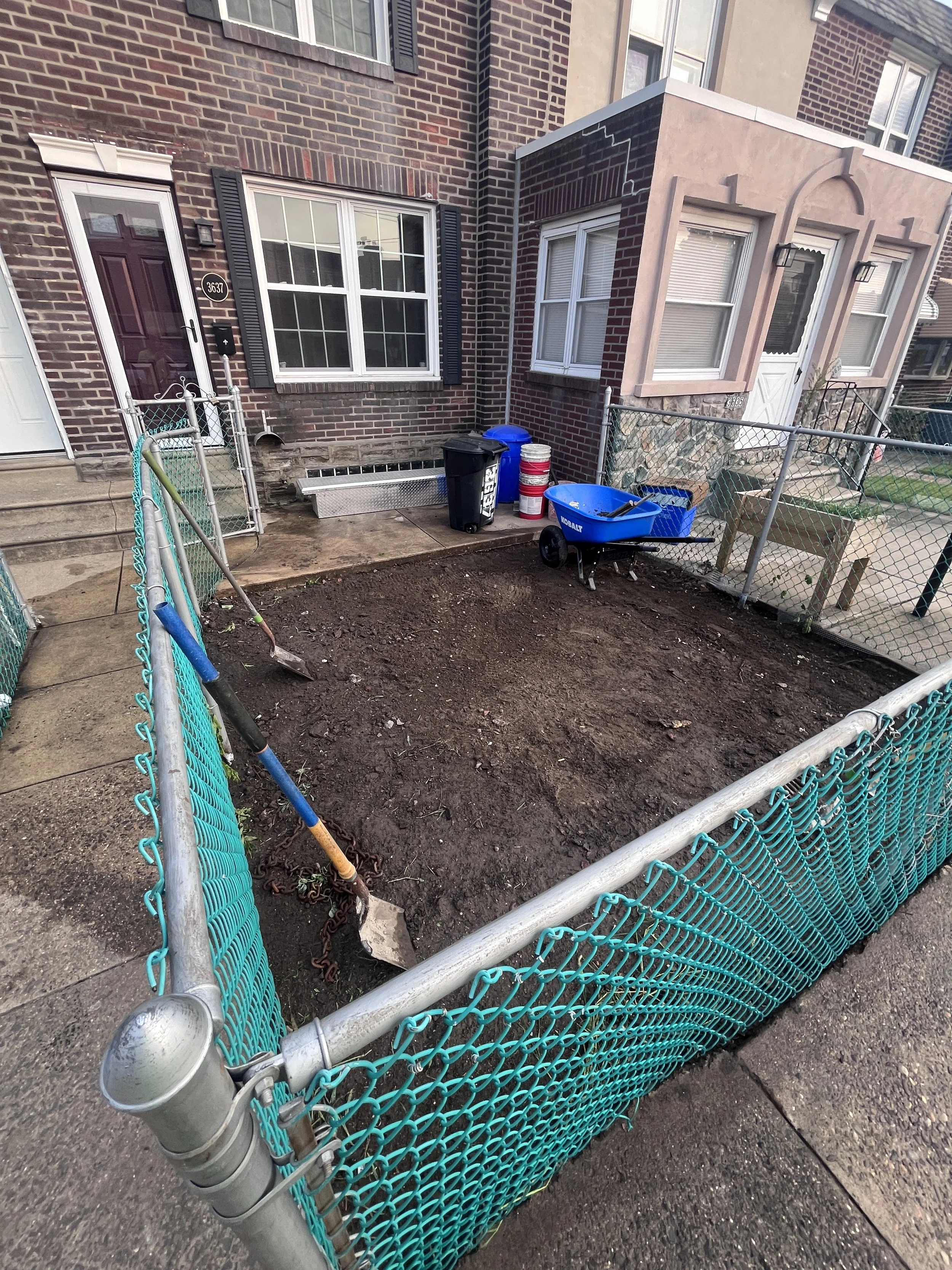 A fenced-off backyard with soil and some gardening tools, including a hand shovel and a yard cart, in front of a brick apartment building with several windows.