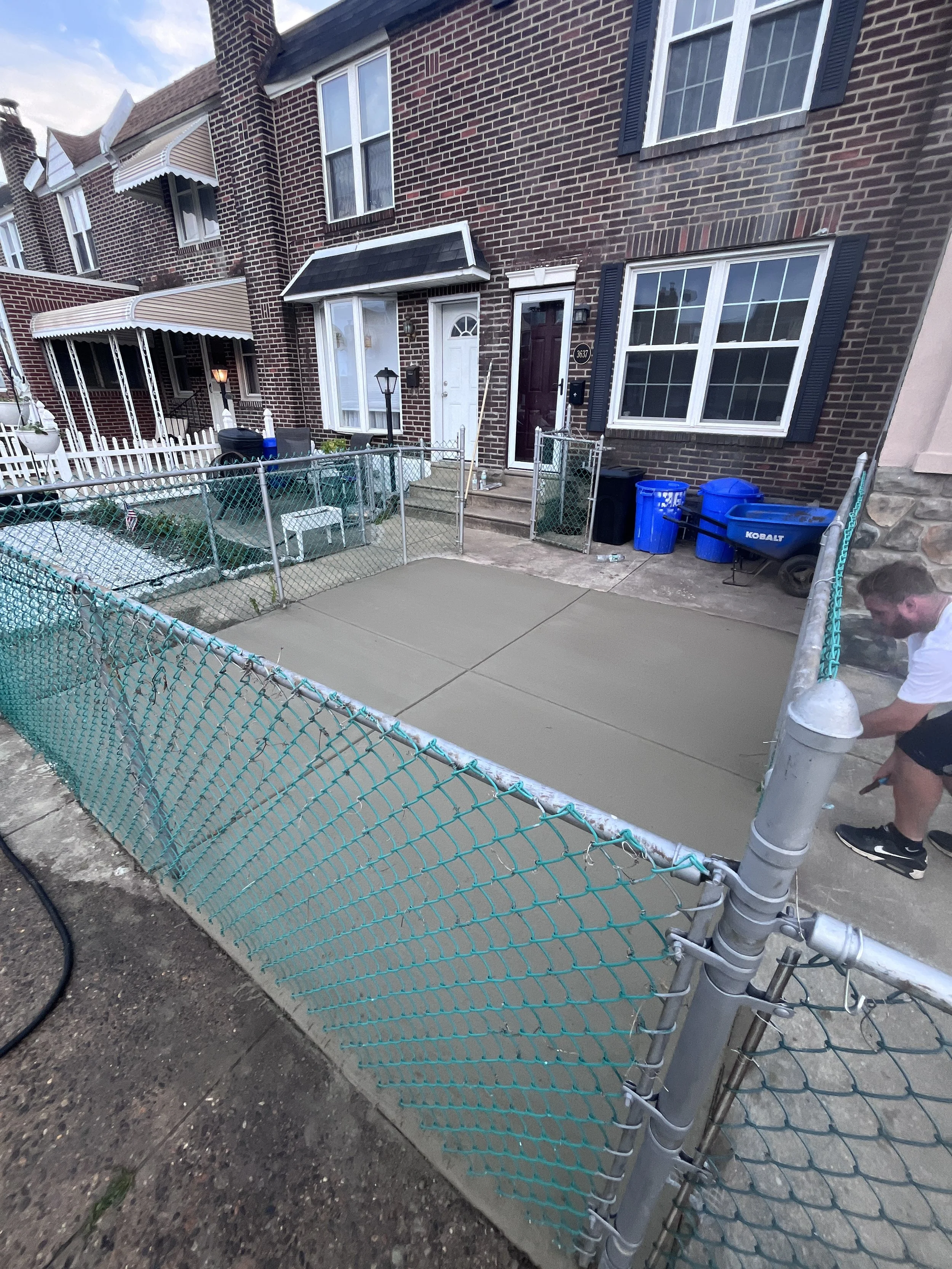 A sidewalk in front of a brick house with a fenced yard. A person is working on the fence, attaching a new section of chain-link. The house has multiple windows, some with black shutters, and an awning over the front door. There are trash cans and a 