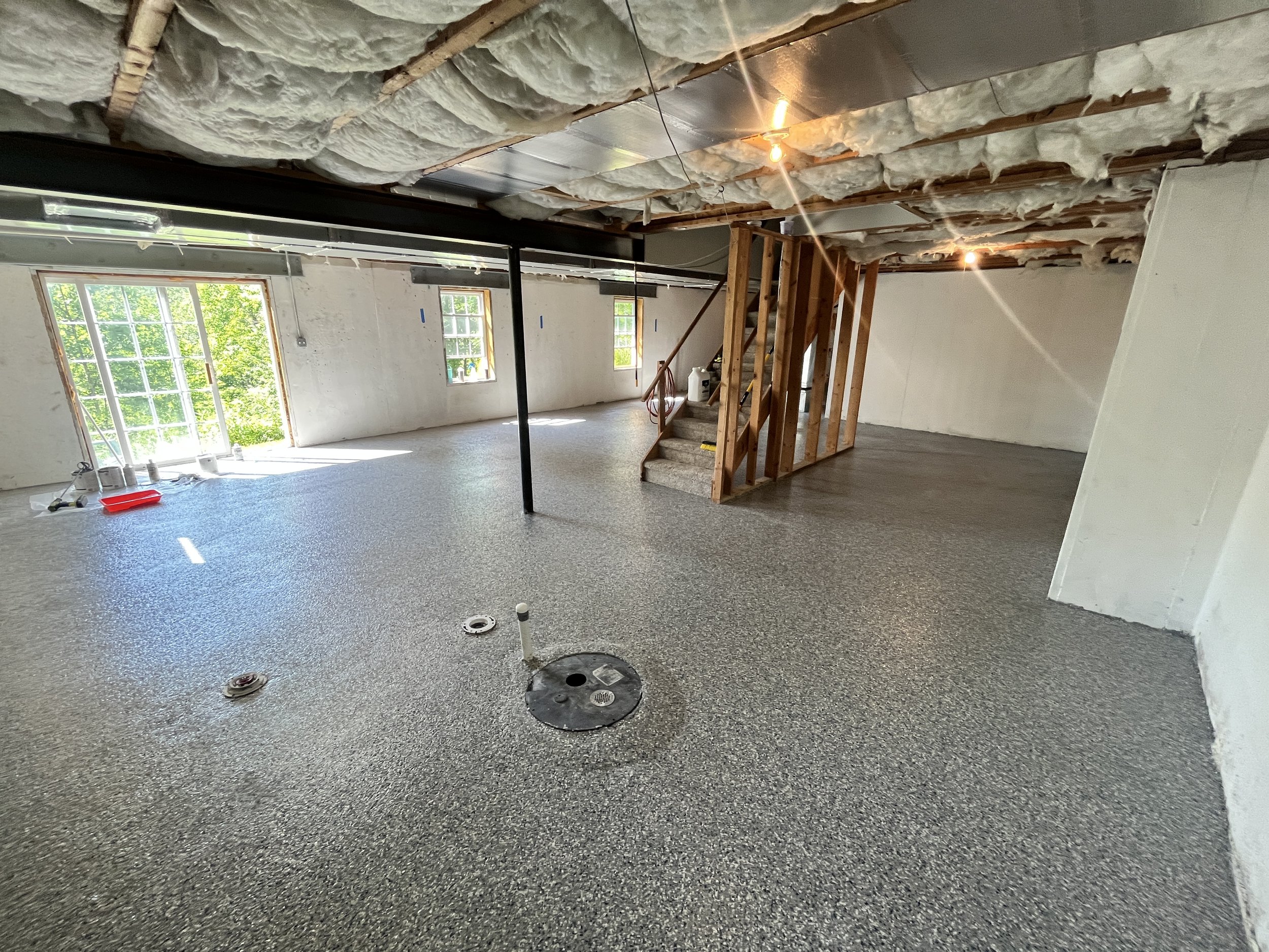 Interior of a house under renovation showing unfinished ceiling with insulation, a black metal support beam, wooden staircase framing, and a window with green foliage outside.