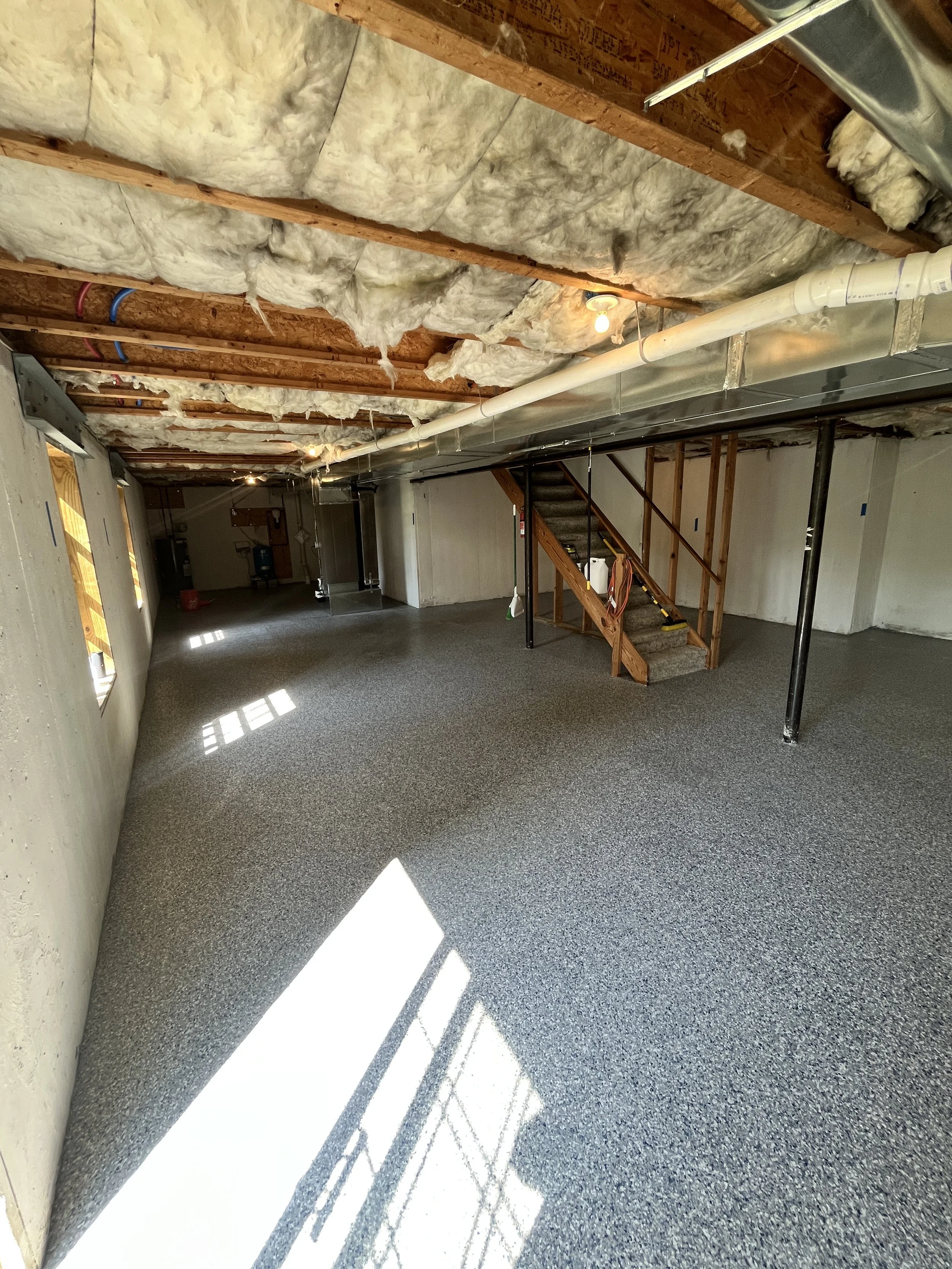 Empty basement room with unfinished ceiling, insulation, exposed pipes, staircase, and carpeted flooring with sunlight through window.