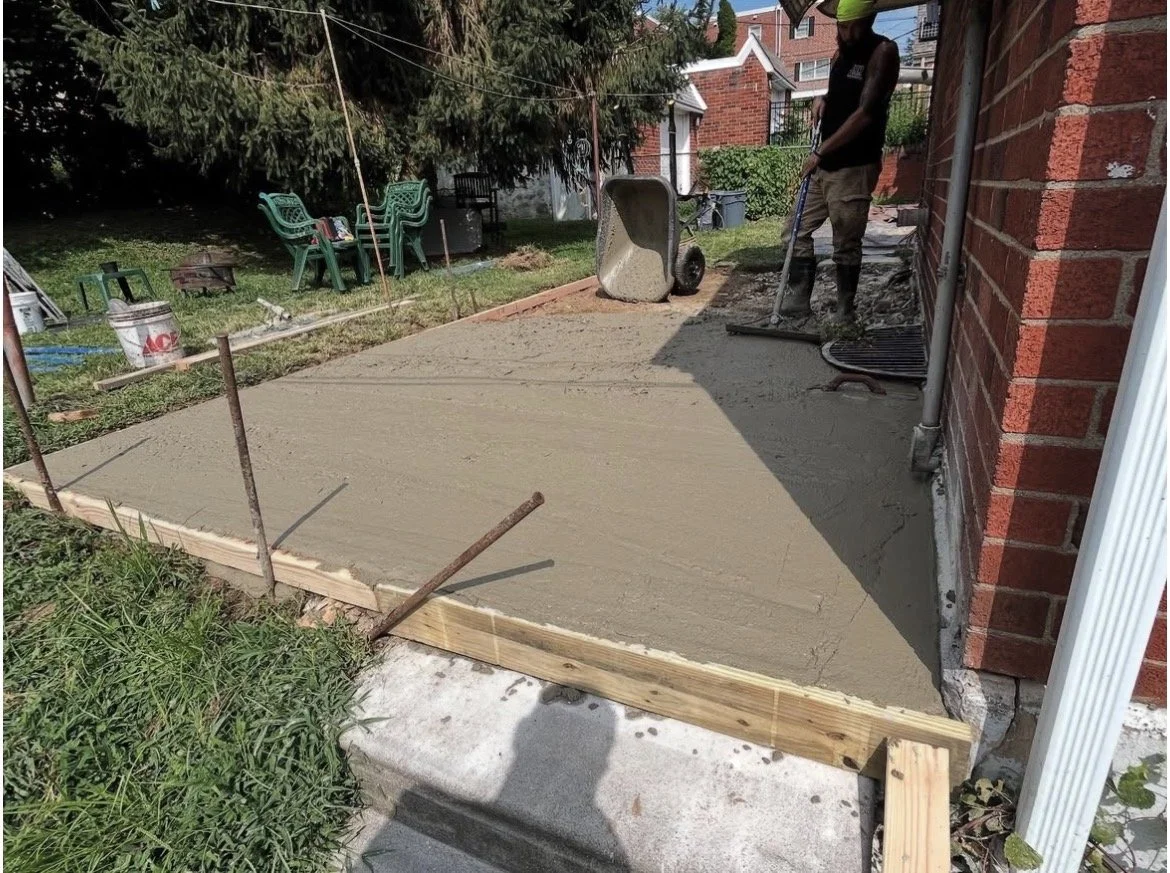 a concrete patio installation outside a brick house, with construction tools and materials nearby.