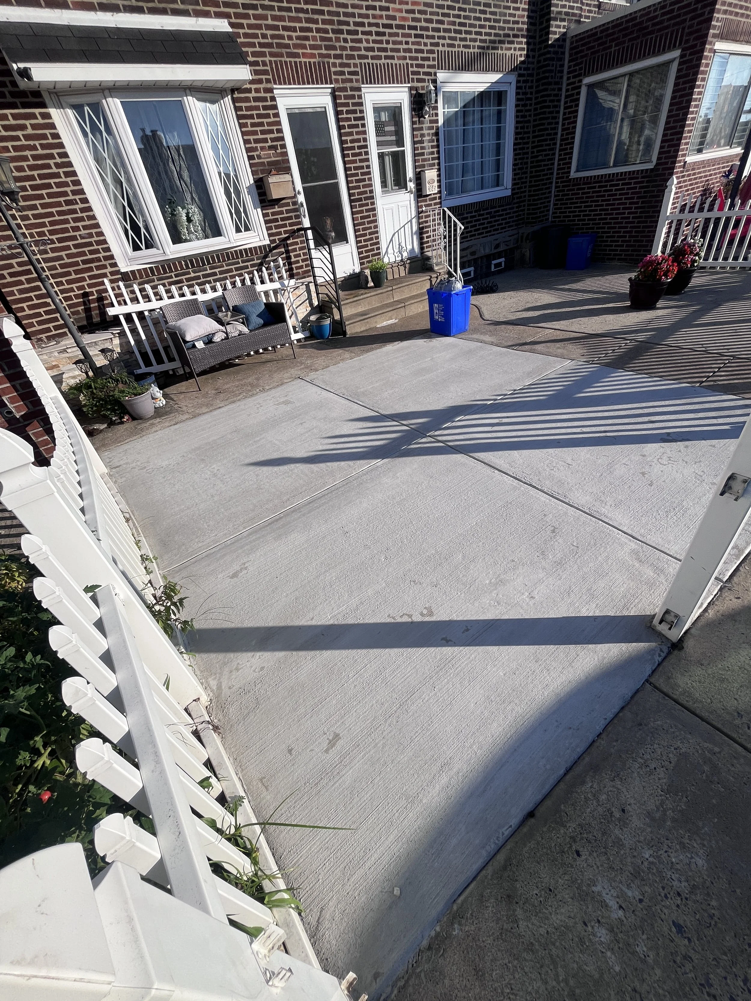 View of a house entrance with brick exterior, windows, white security door, potted plants, a bench, trash bins, and a concrete driveway with shadows.