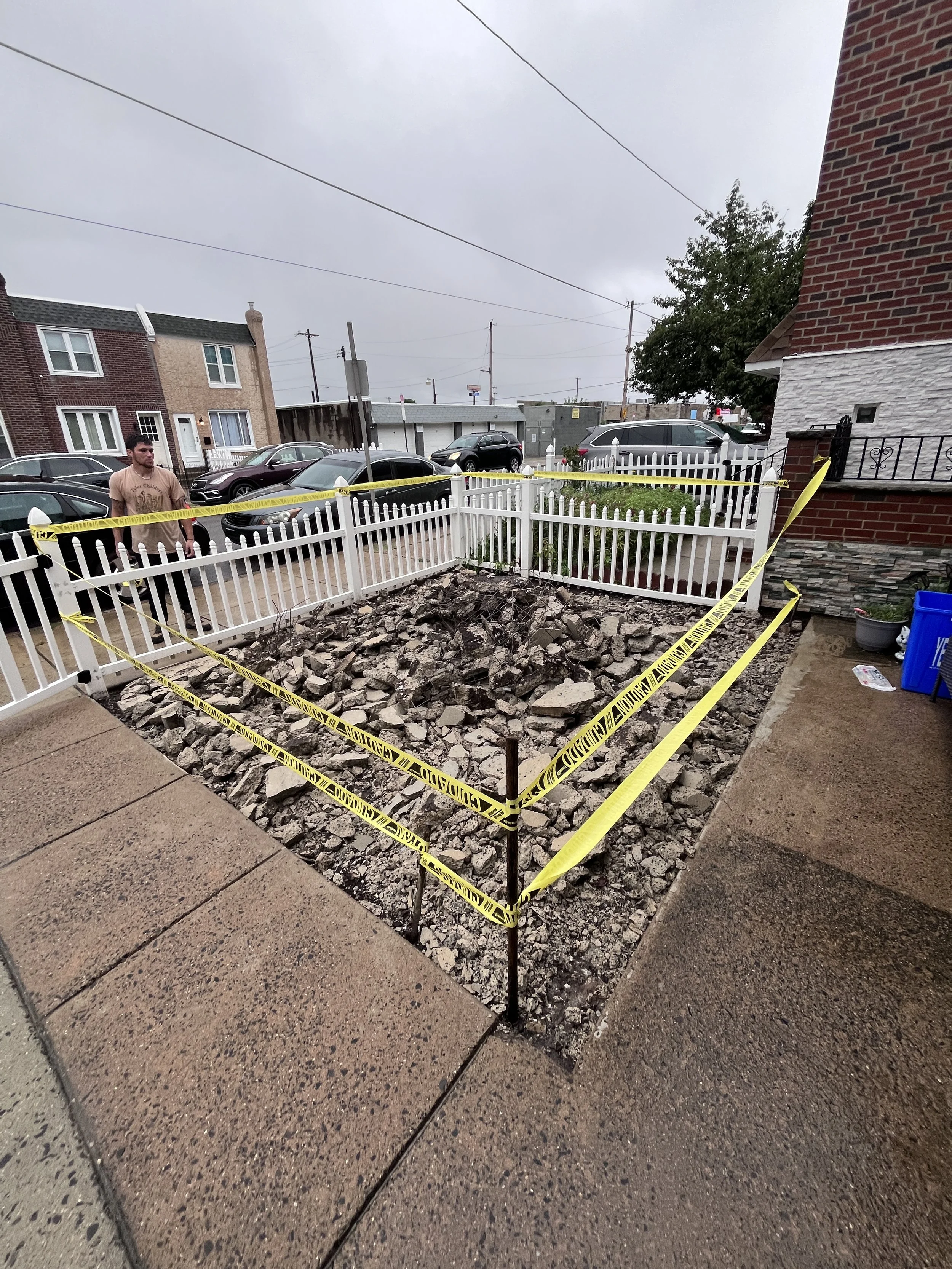 A small fenced yard with a damaged section filled with rocks, surrounded by yellow caution tape, in front of residential buildings and parked cars on a cloudy day.
